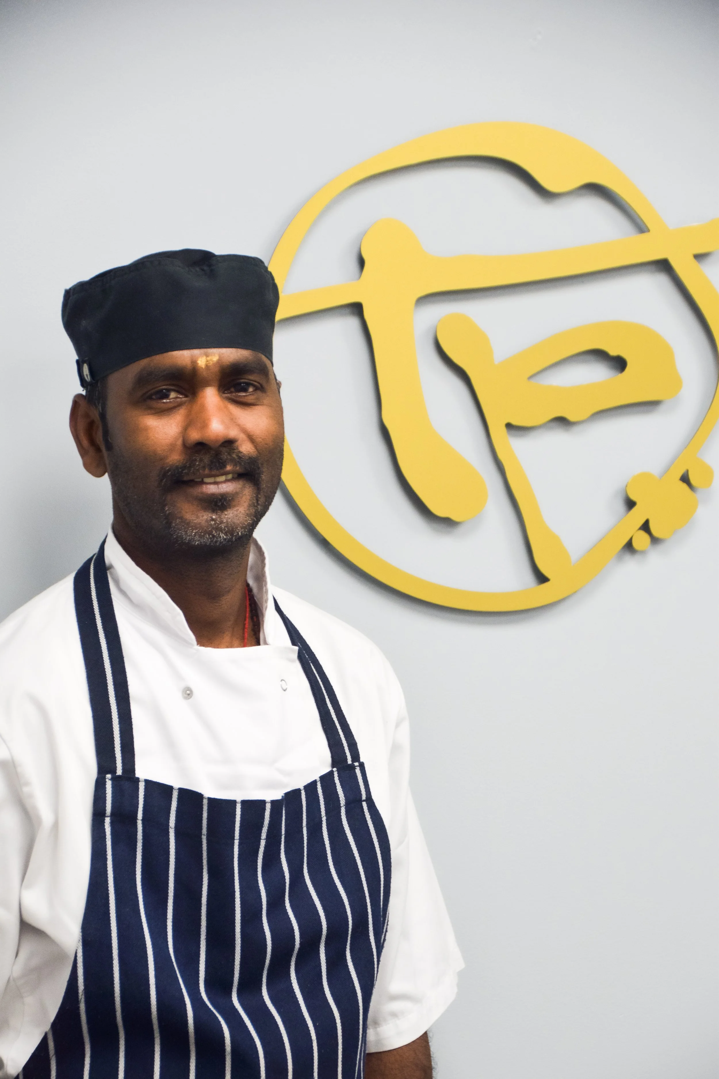 A man in a white chef's coat and black hat standing in front of a wall with a sign that reads "Terry's Batisserie" and a logo featuring a stylized face wearing glasses and a chef's hat.
