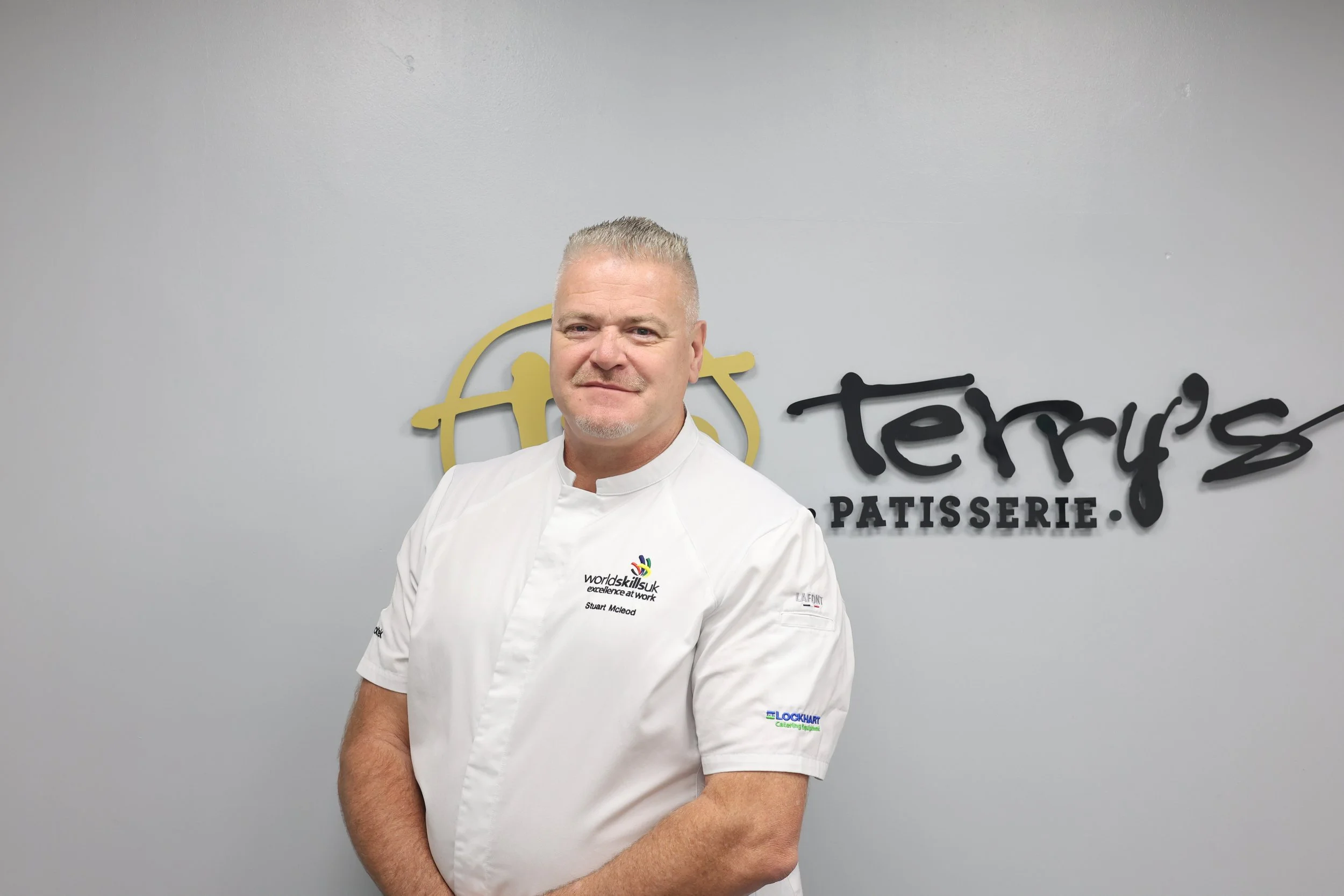 A young boy wearing a plastic apron standing in front of a wall sign that reads 'terry's Patisserie'.