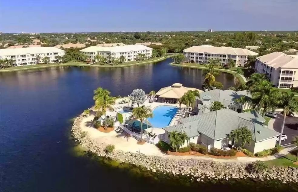 Aerial view of a residential community featuring a central lake, a swimming pool, surrounding buildings, and palm trees.