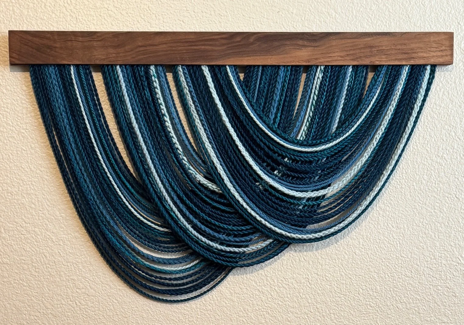 Wall hanging art piece made of multiple blue, white, black, and teal yarn strands draped from a wooden rod mounted on a wall.