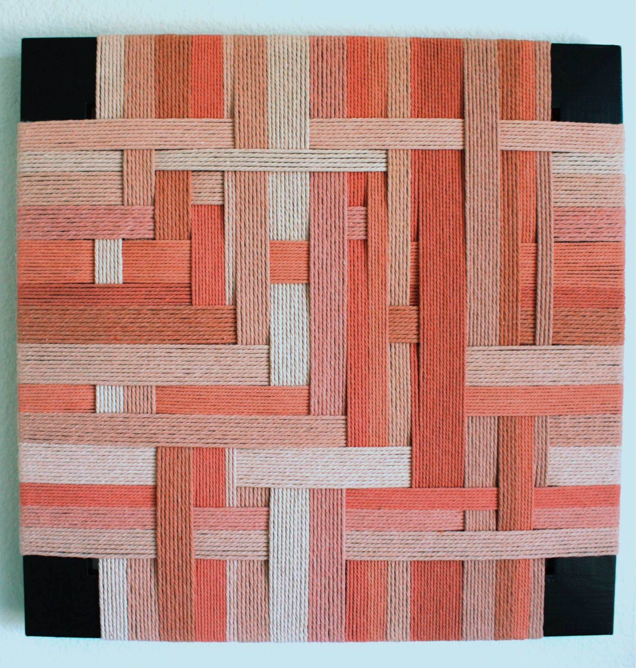 A woven basket pattern made of interlaced yarn in shades of pink, coral, and beige with black borders at the corners.