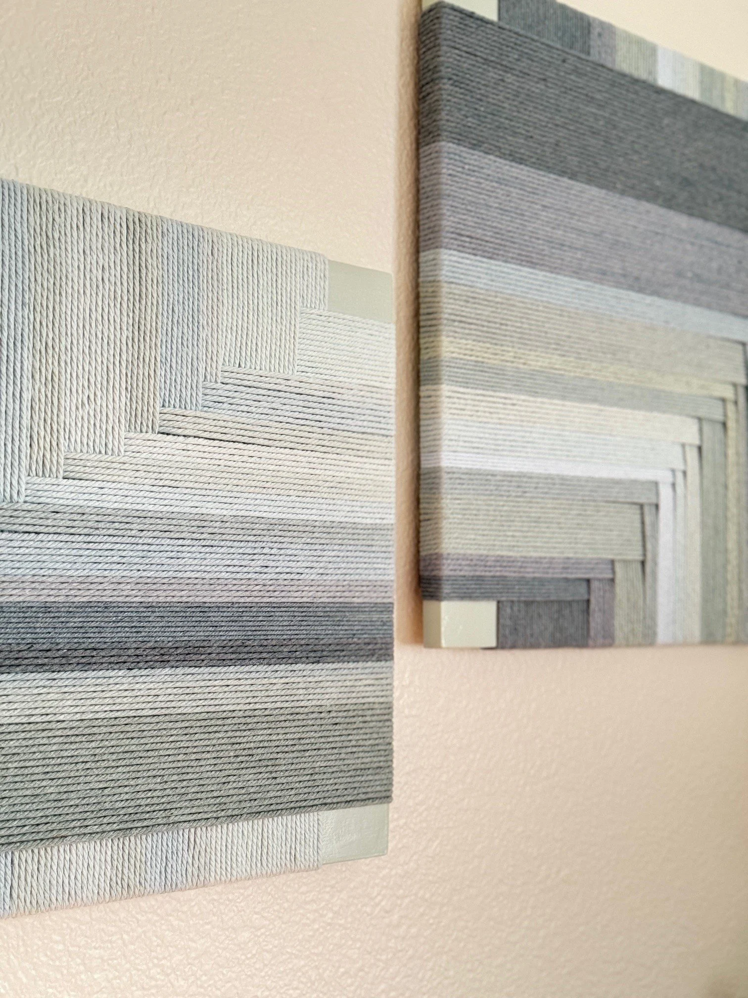 Close-up of two abstract textured canvas artworks with geometric patterns in shades of gray and white, mounted on a pink wall.