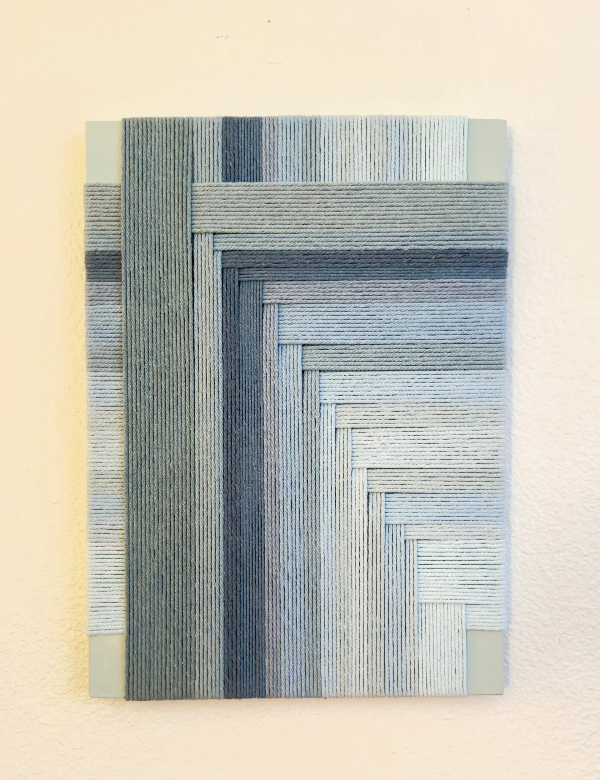 Wall art featuring woven fabric strips in shades of gray, arranged in a geometric pattern on a white background.