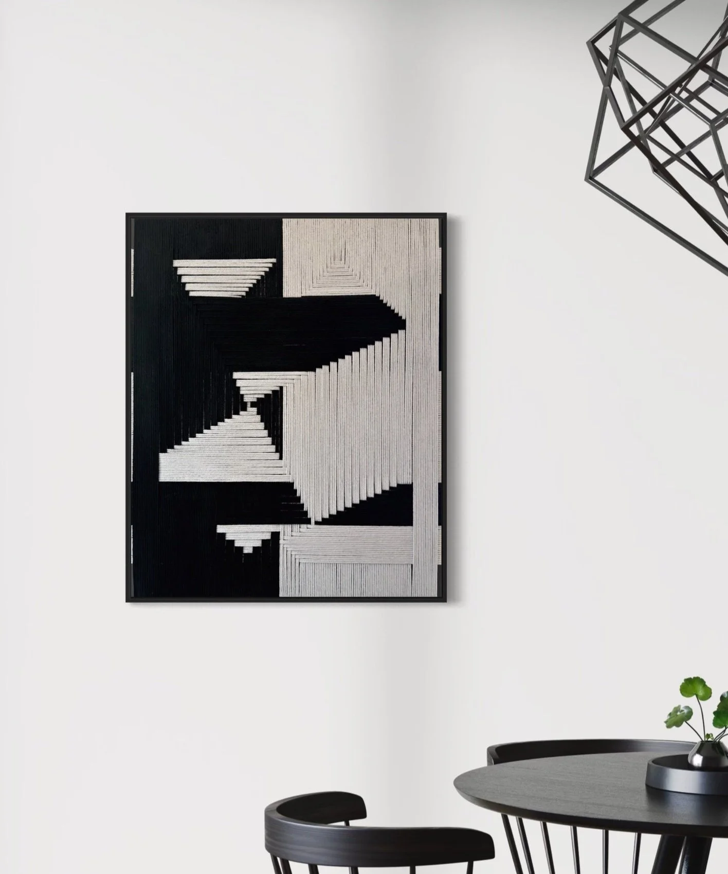 Modern black and white geometric art print hanging on a white wall in a minimalistic dining area with a round table and black chairs, a small plant, and a geometric wall sculpture.