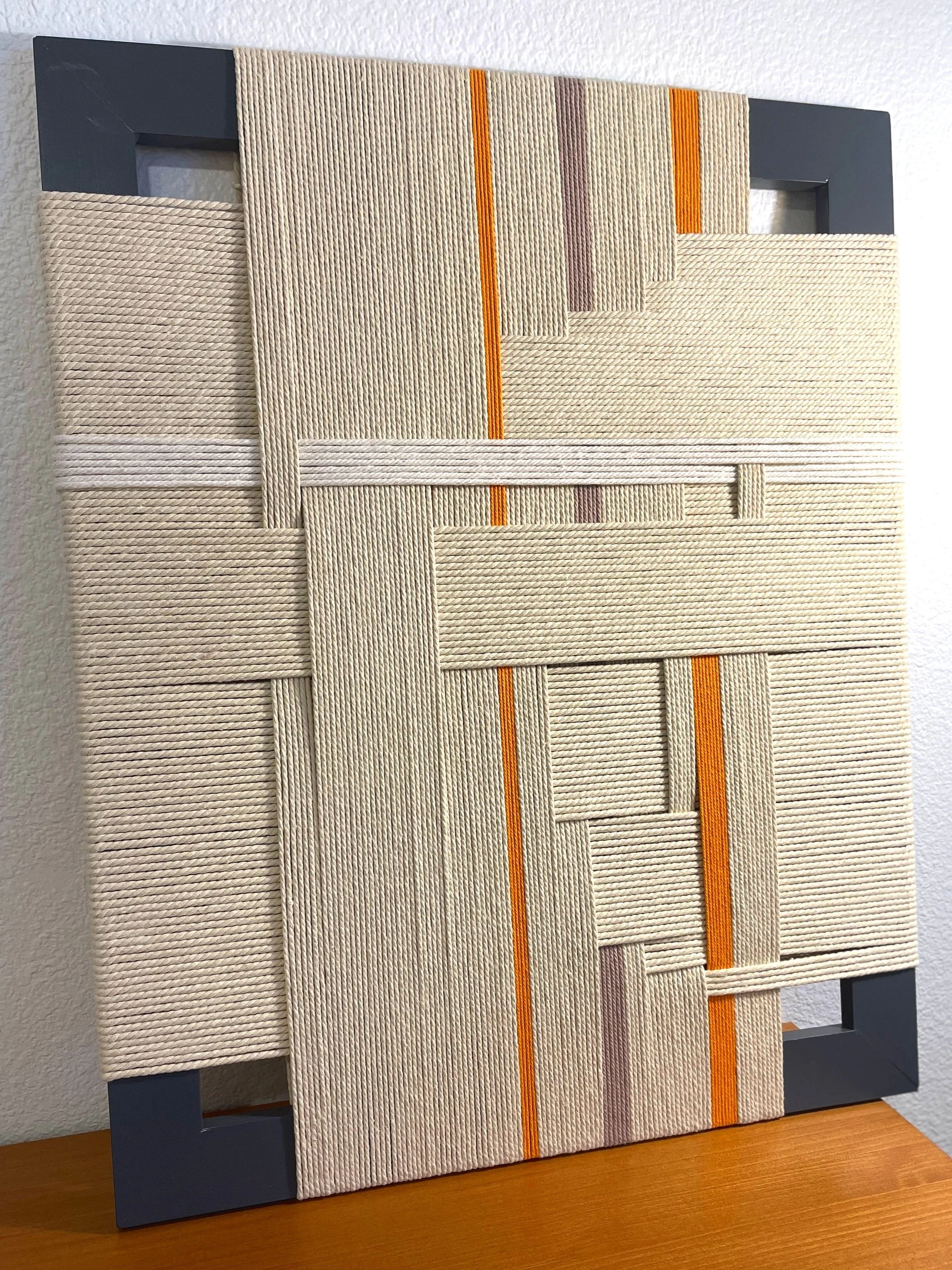 A modern woven wall art piece with a geometric pattern, featuring beige, cream, purple, orange, and gray strips, mounted on a black frame.