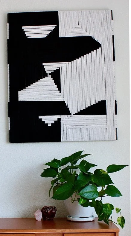 Black and white geometric wall art with layered rectangular patterns, mounted above a wooden surface with a green potted plant, a small carved object, and a pink rock.