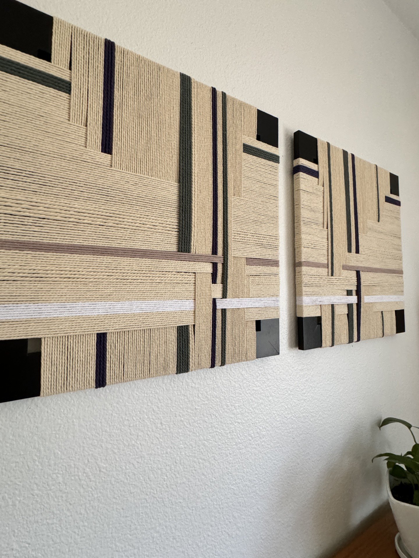 Two abstract wall art pieces with geometric pattern and textured fabric on a white wall, with a small plant in a white pot below.