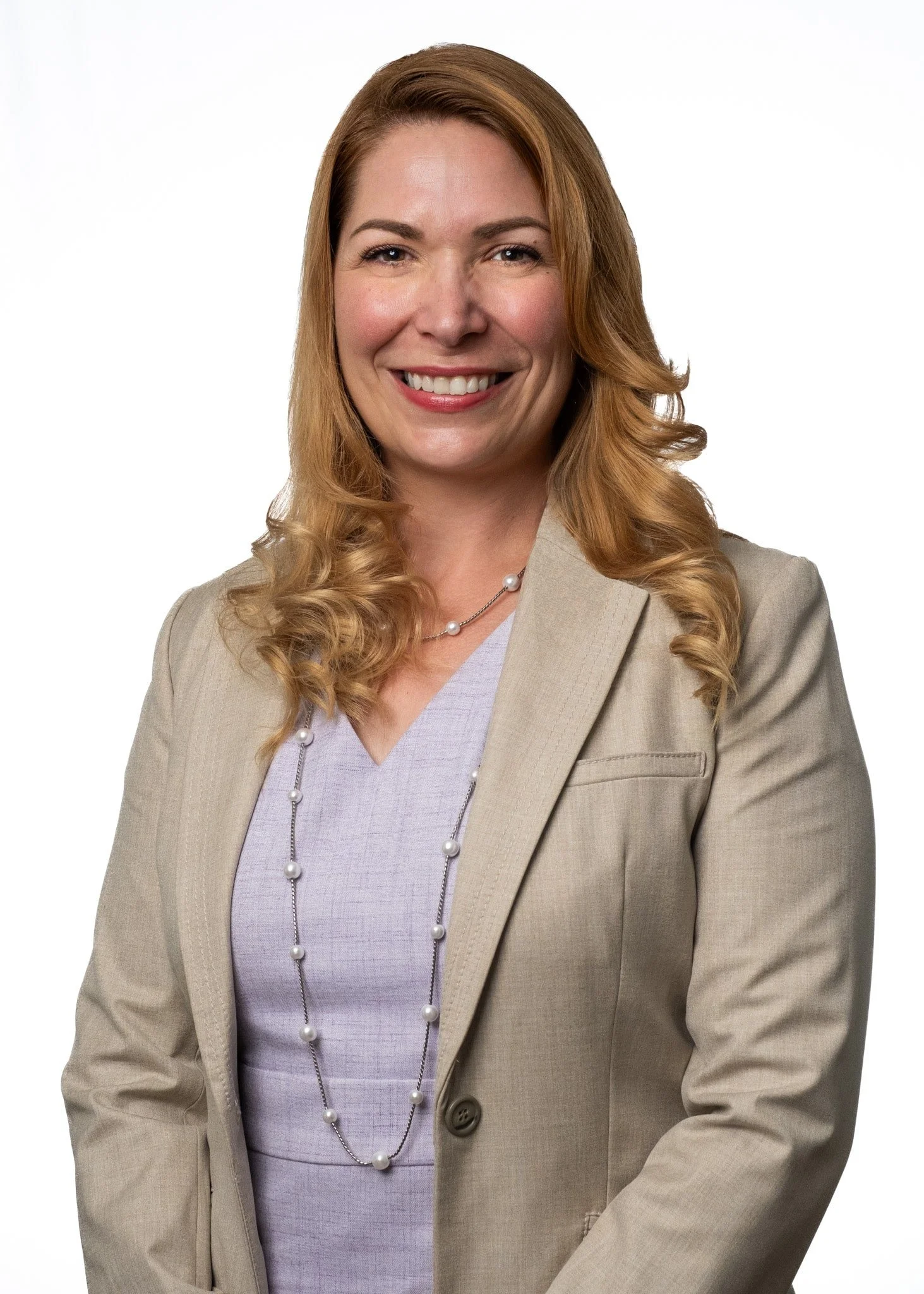 A smiling person with wavy, shoulder-length blonde hair wearing a beige blazer and a lilac top, accessorized with a long pearl necklace, standing against a plain white background.