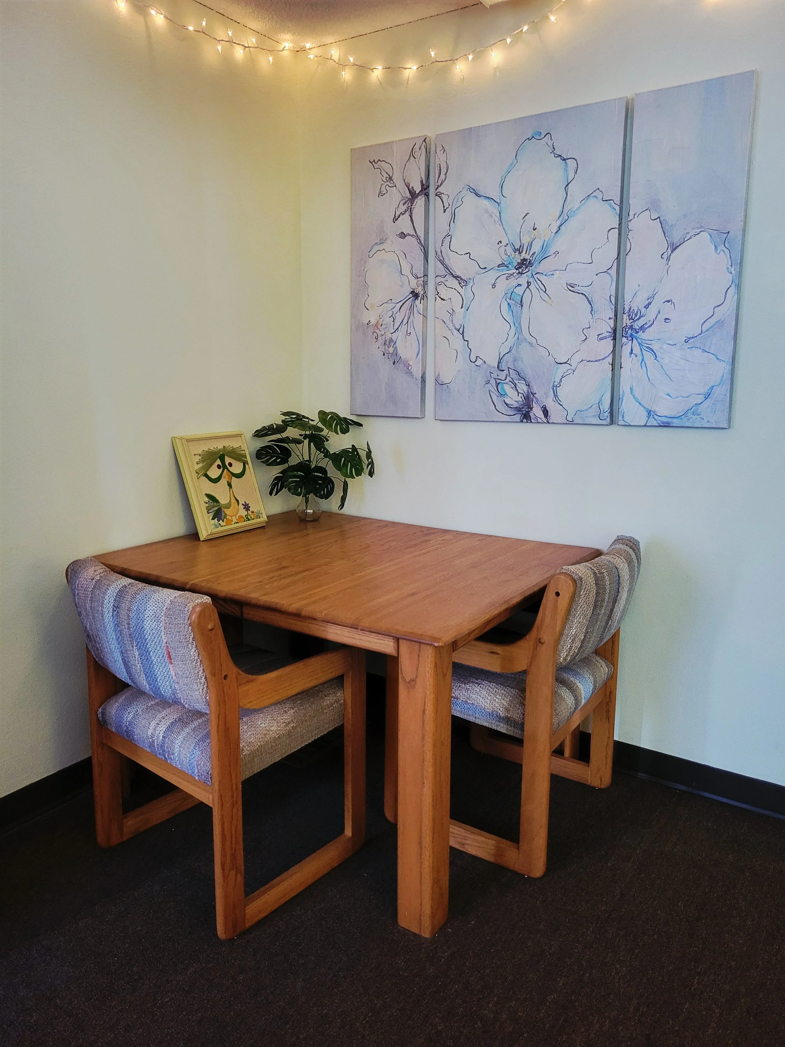 Corner of a room with a wooden table and two upholstered chairs, a framed picture of a stylized peacock, a potted plant, and a large floral painting on the wall, with string lights hanging overhead.