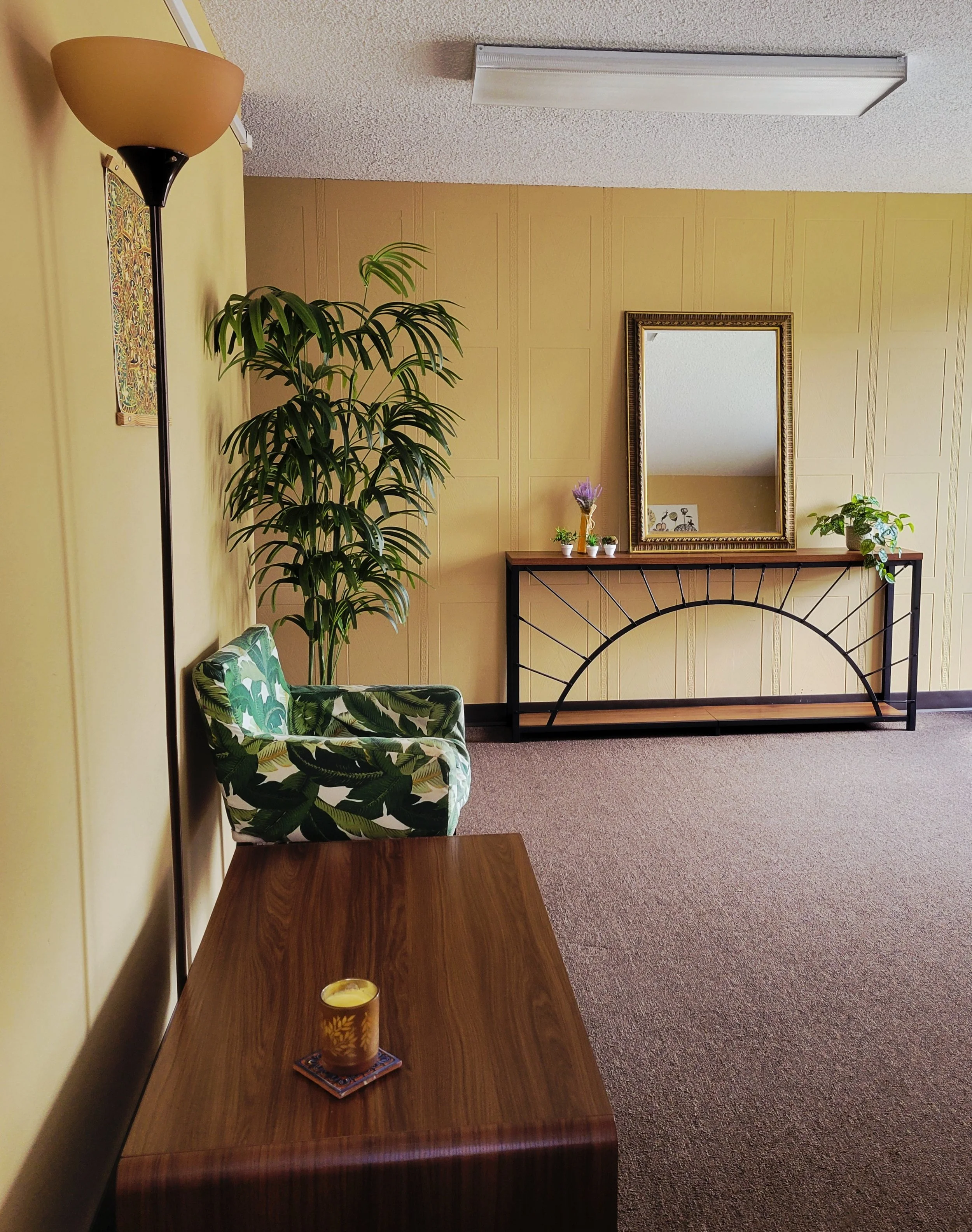 A cozy living room corner with a wooden table, a patterned armchair with green leaf design, a tall potted plant, a mirror on a narrow black metal and wood console table, and decorative plants and flowers on the table. The room has yellow walls and brown carpeting.