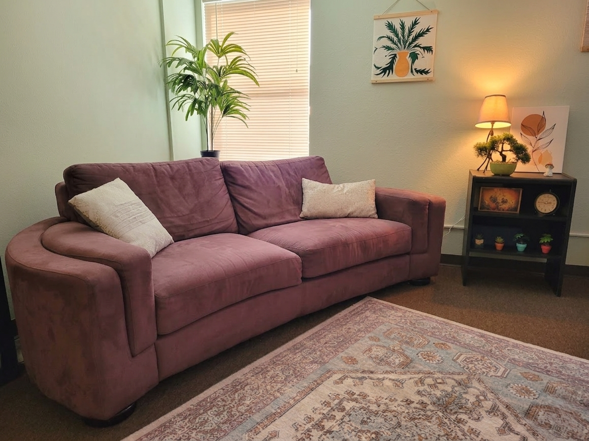 Living room with a purple sofa, two white pillows, potted plant, wall art, side table with a lamp, small bonsai, and decorative plants.