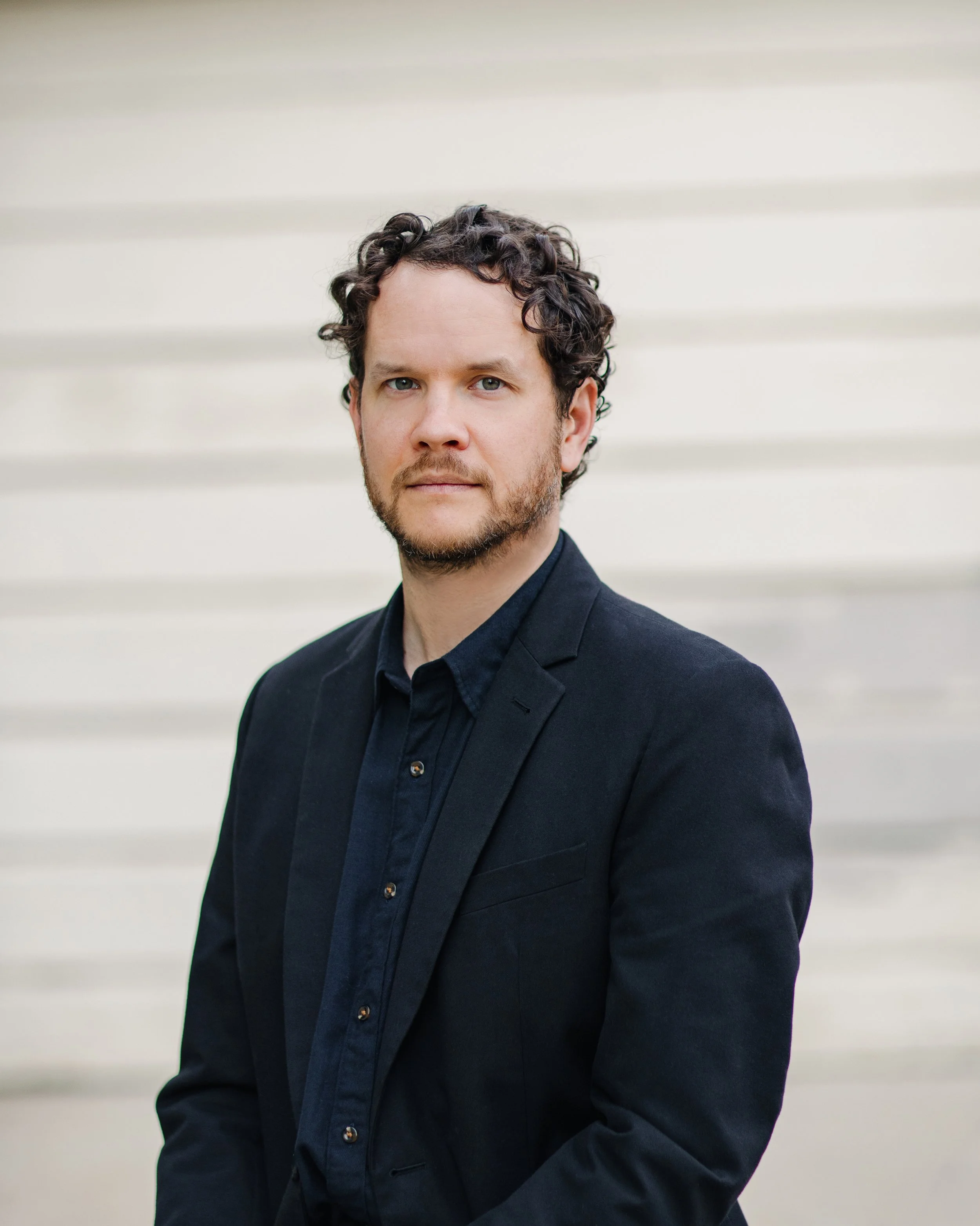 A man with curly dark hair and light stubble is wearing a black suit jacket and a dark shirt, standing against a light-colored horizontal siding wall.