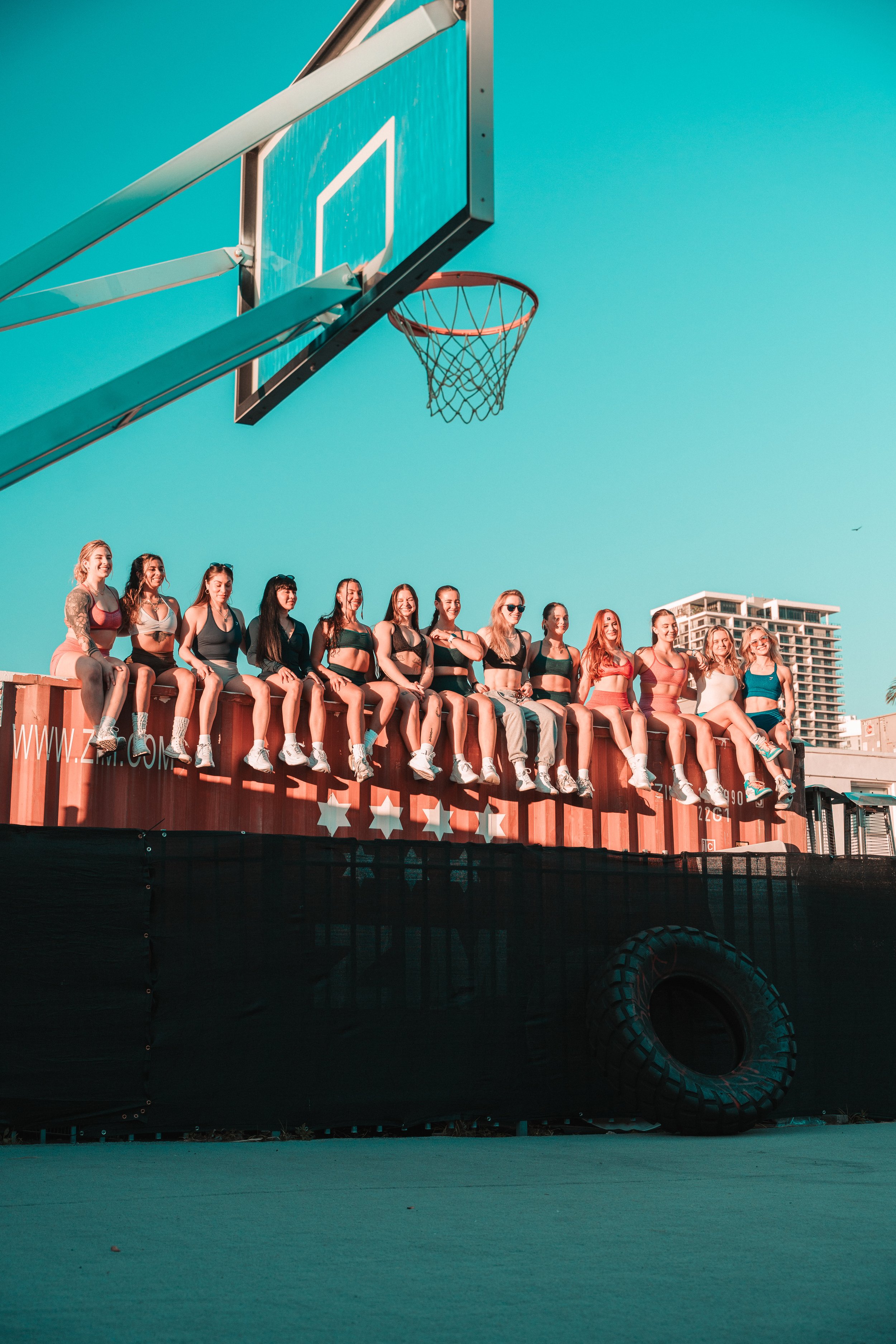 Group of women dressed in athletic wear sitting on a large black platform with red, white, and blue star decorations, beside a basketball hoop and backboard during daytime with a clear blue sky.
