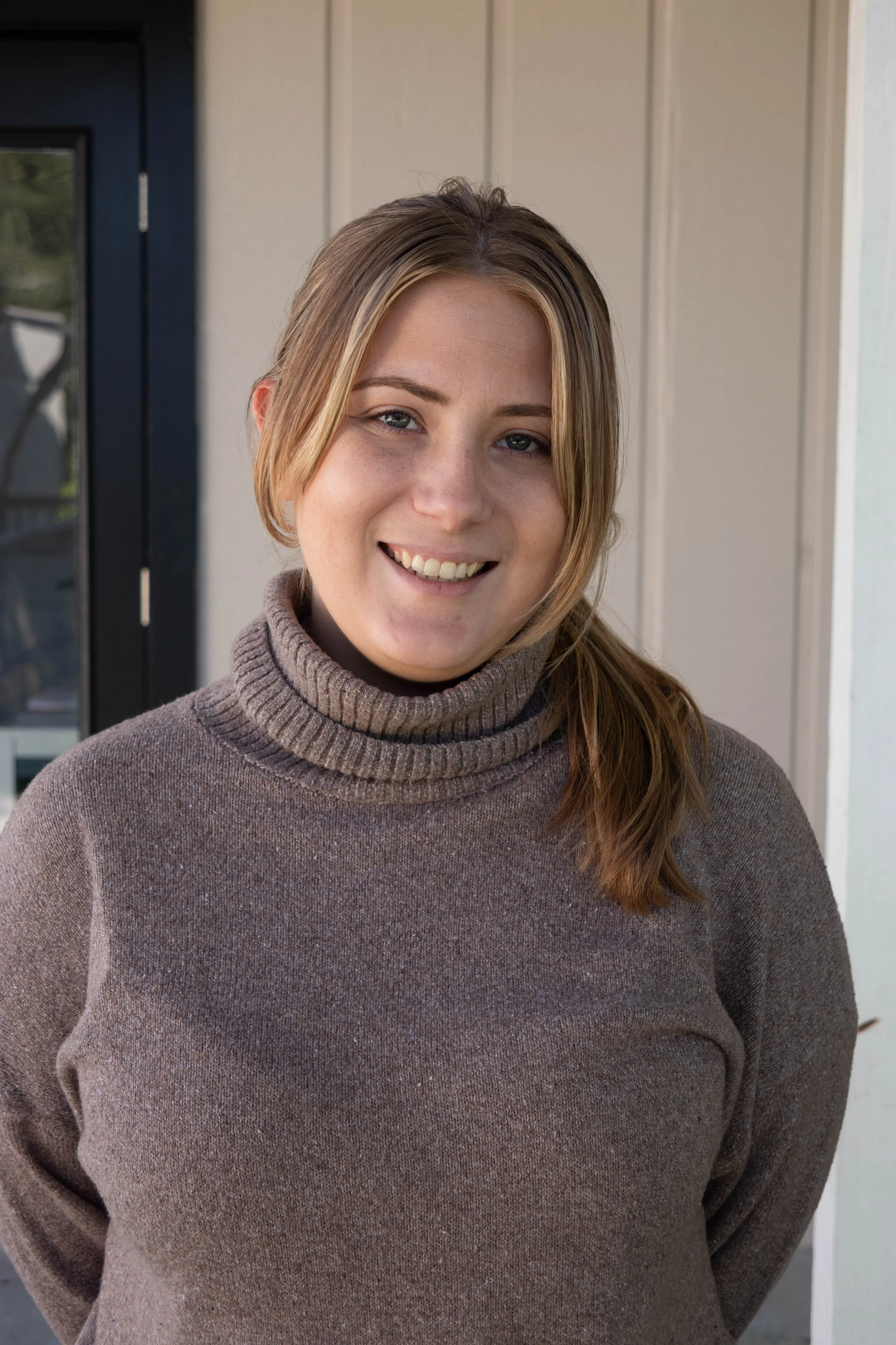 A woman with light brown hair in a ponytail, smiling in front of a beige wall with a black door on her left.
