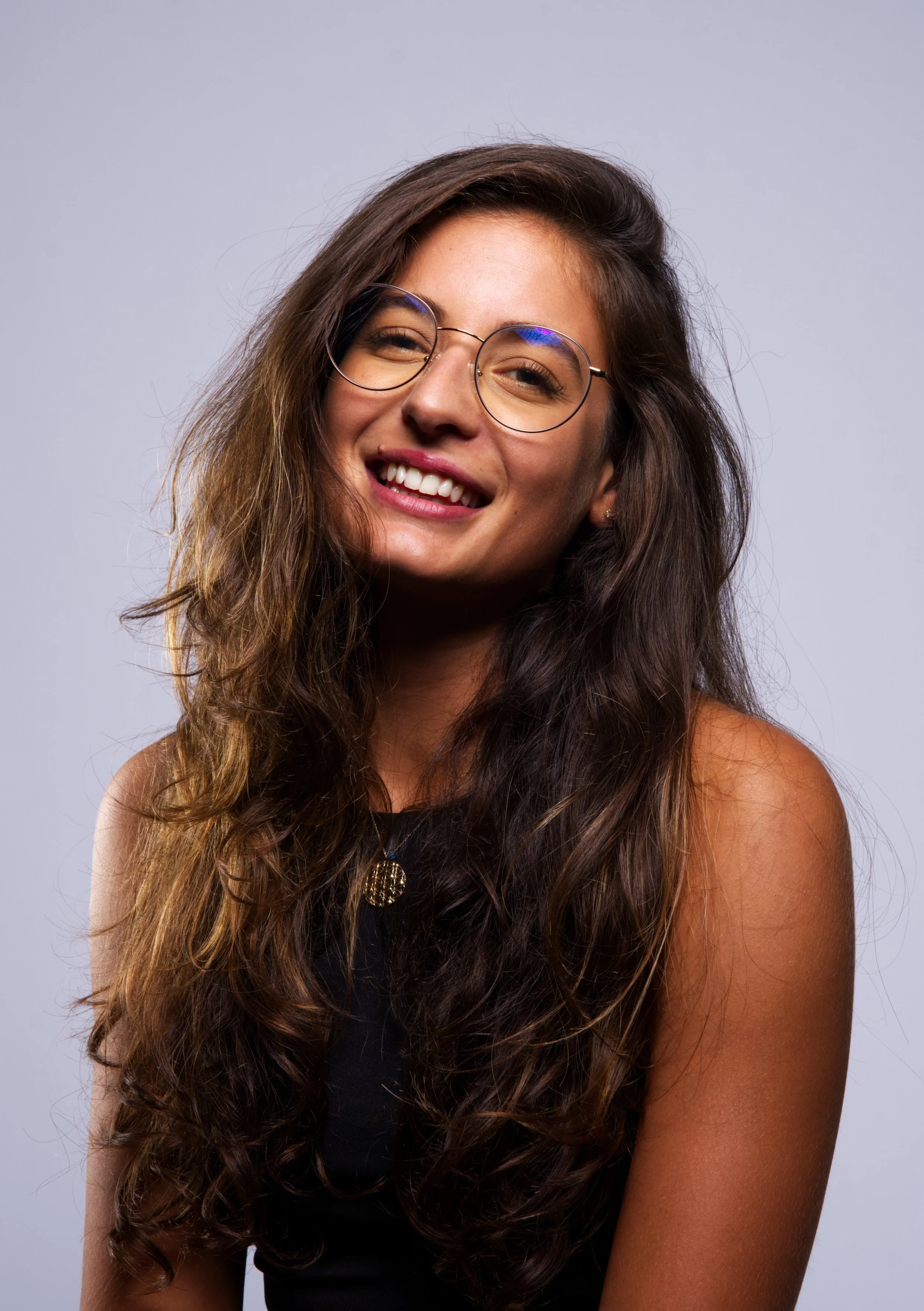 Frau mit langen braunen Haaren und Brille, lächelnd, vor grauem Hintergrund.