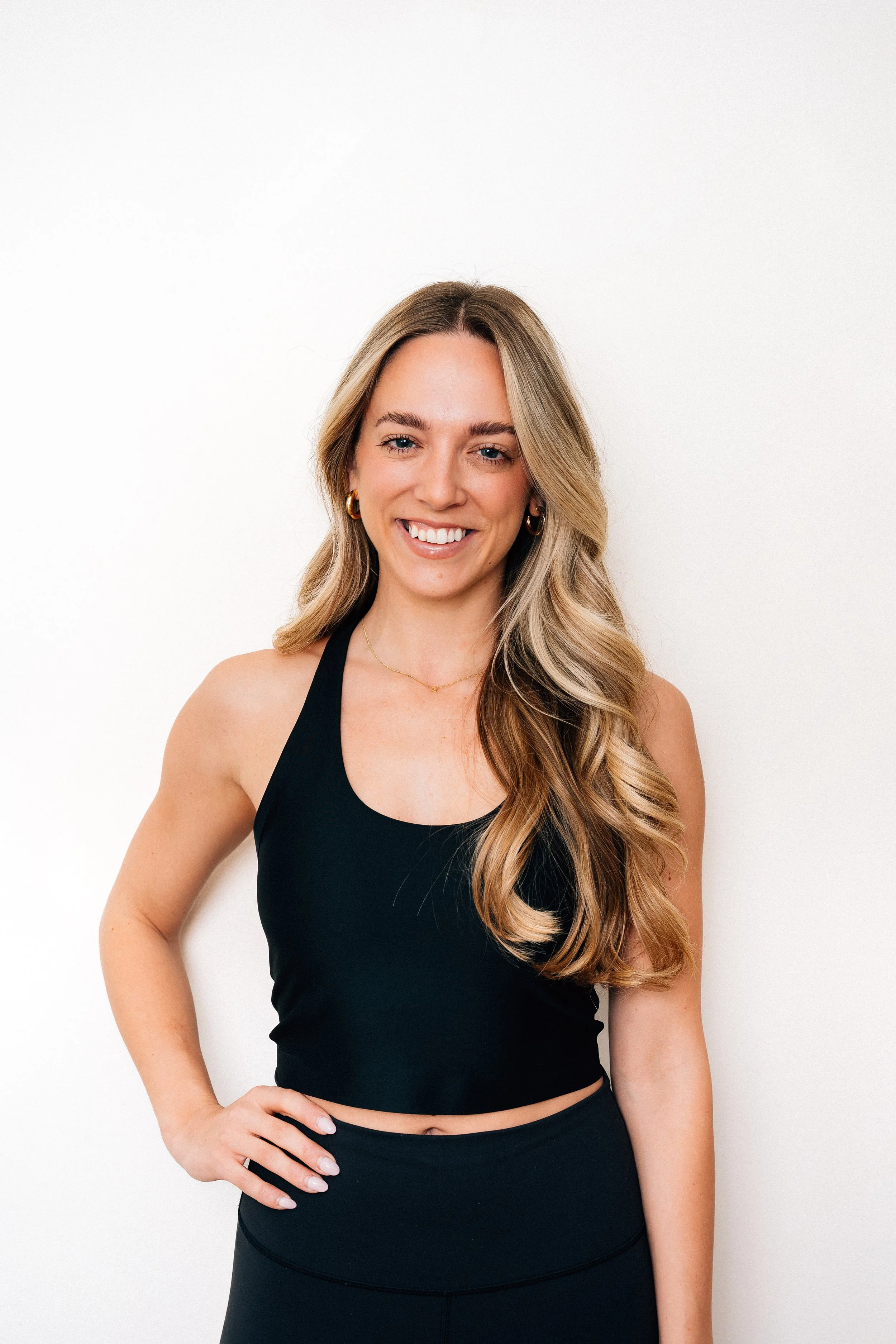 A woman with long, wavy blonde hair smiling and standing with her hand on her hip, wearing a black sports bra and black leggings, against a plain white background.
