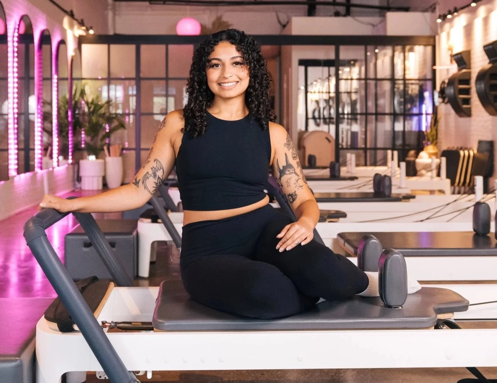 A woman with curly black hair, tattoos on her arms, wearing a black sleeveless top and black pants, sitting on a Pilates reformer machine in a modern gym with pink and purple lighting.