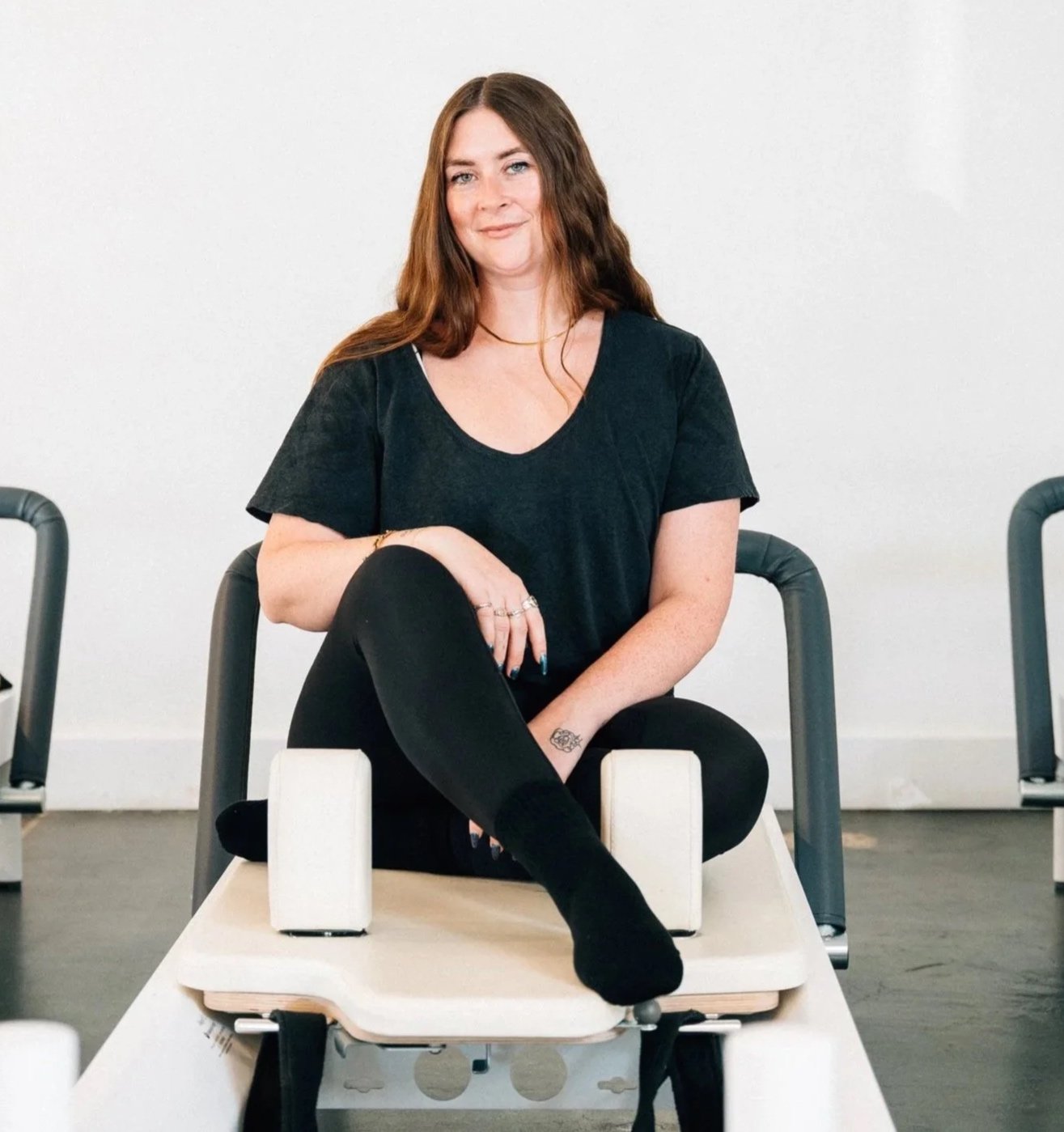 Woman sitting on a chair with one leg crossed over the other, smiling, in a minimalistic room.