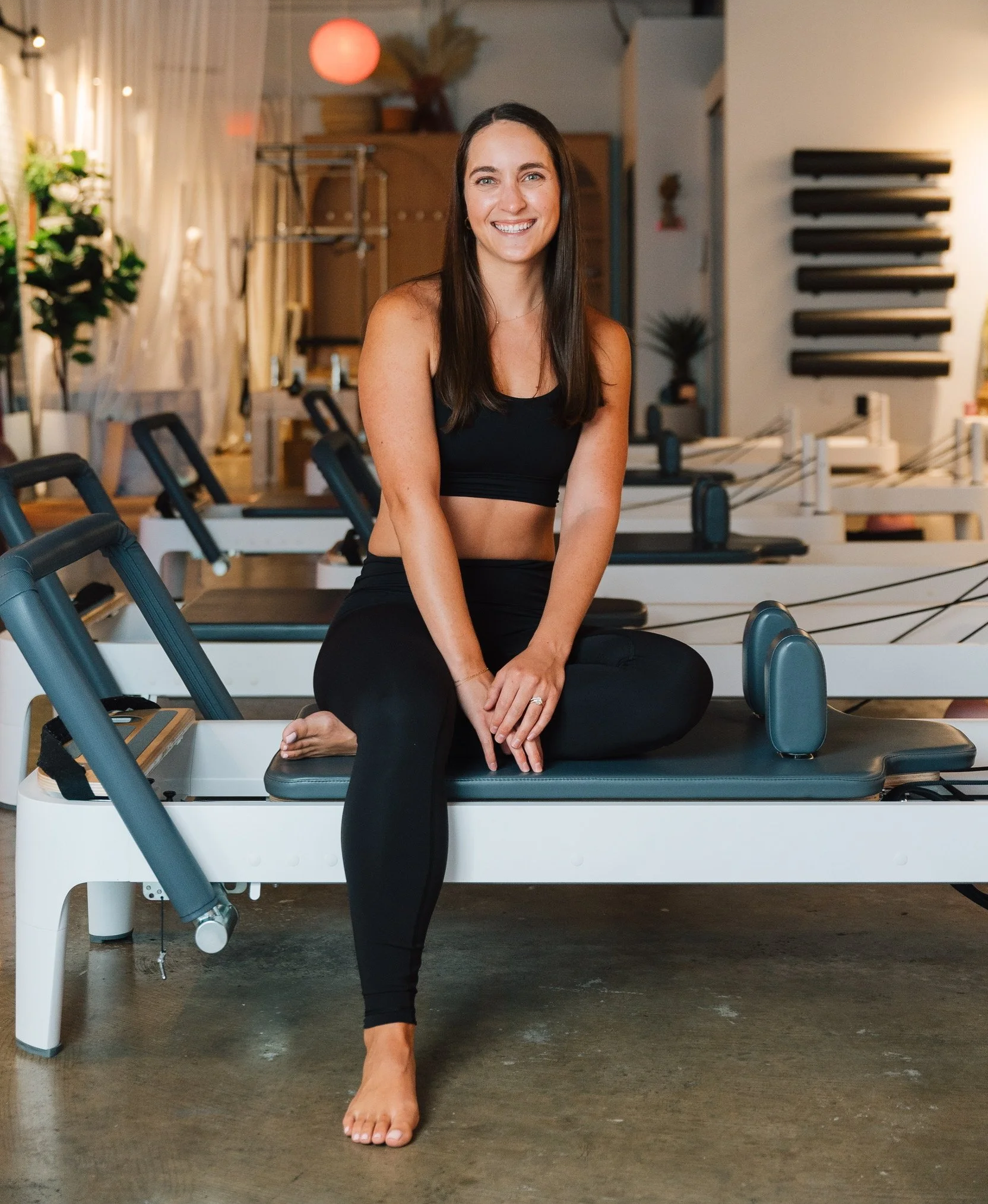 A woman in black workout wear sitting on a Pilates reformer machine in a fitness studio, smiling happily.