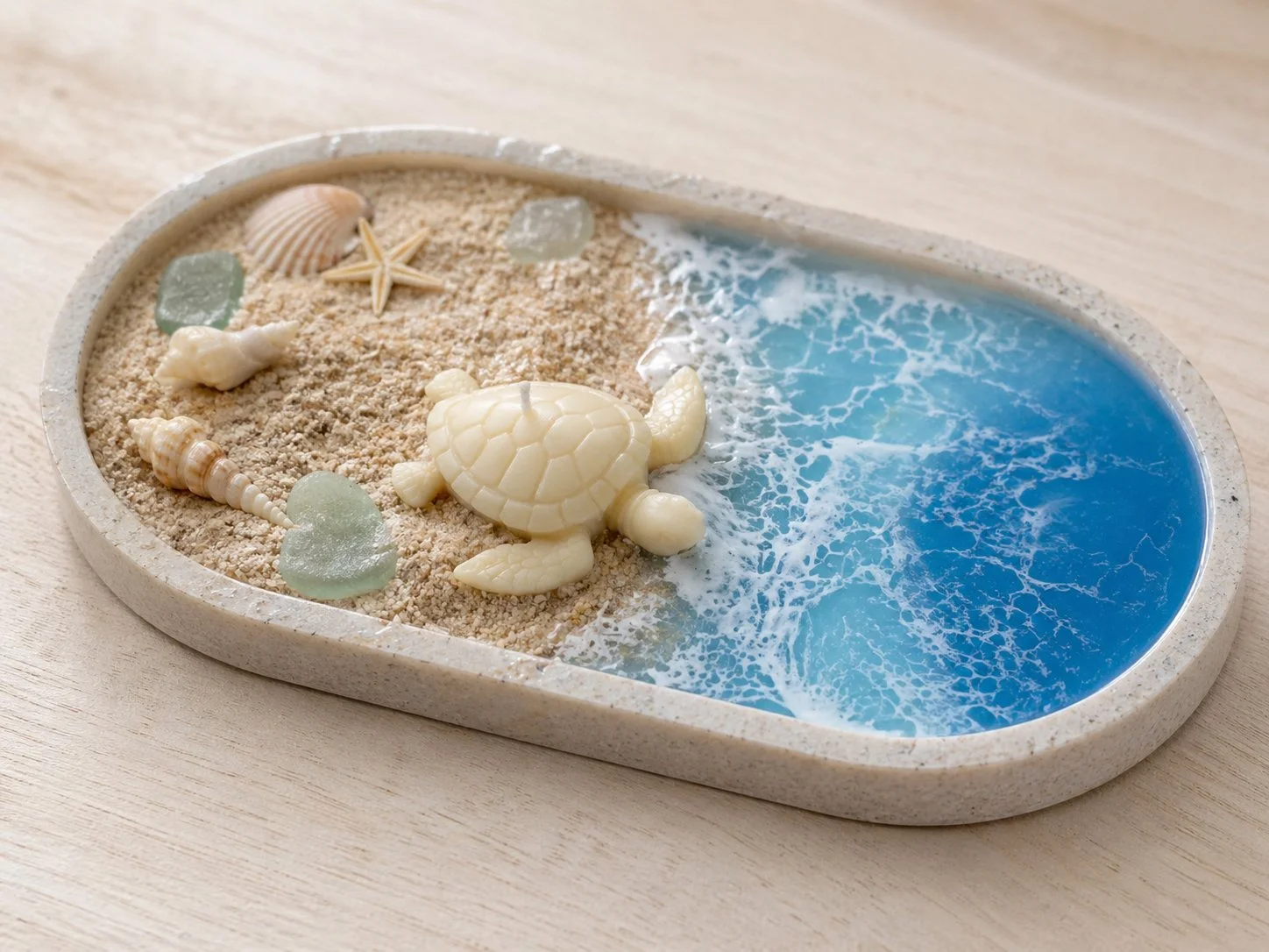 A decorative oval tray with a sandy beach scene, including seashells, glass stones, a starfish, and a small turtle figurine, alongside a blue ocean scene with waves.