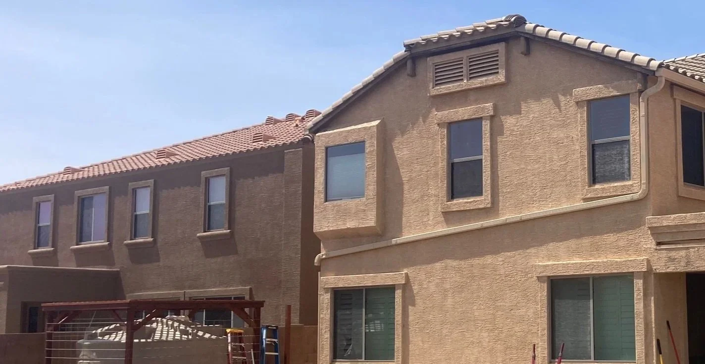 Backyard of a beige stucco house under construction with a woman standing in the foreground, construction tools, and materials scattered around.
