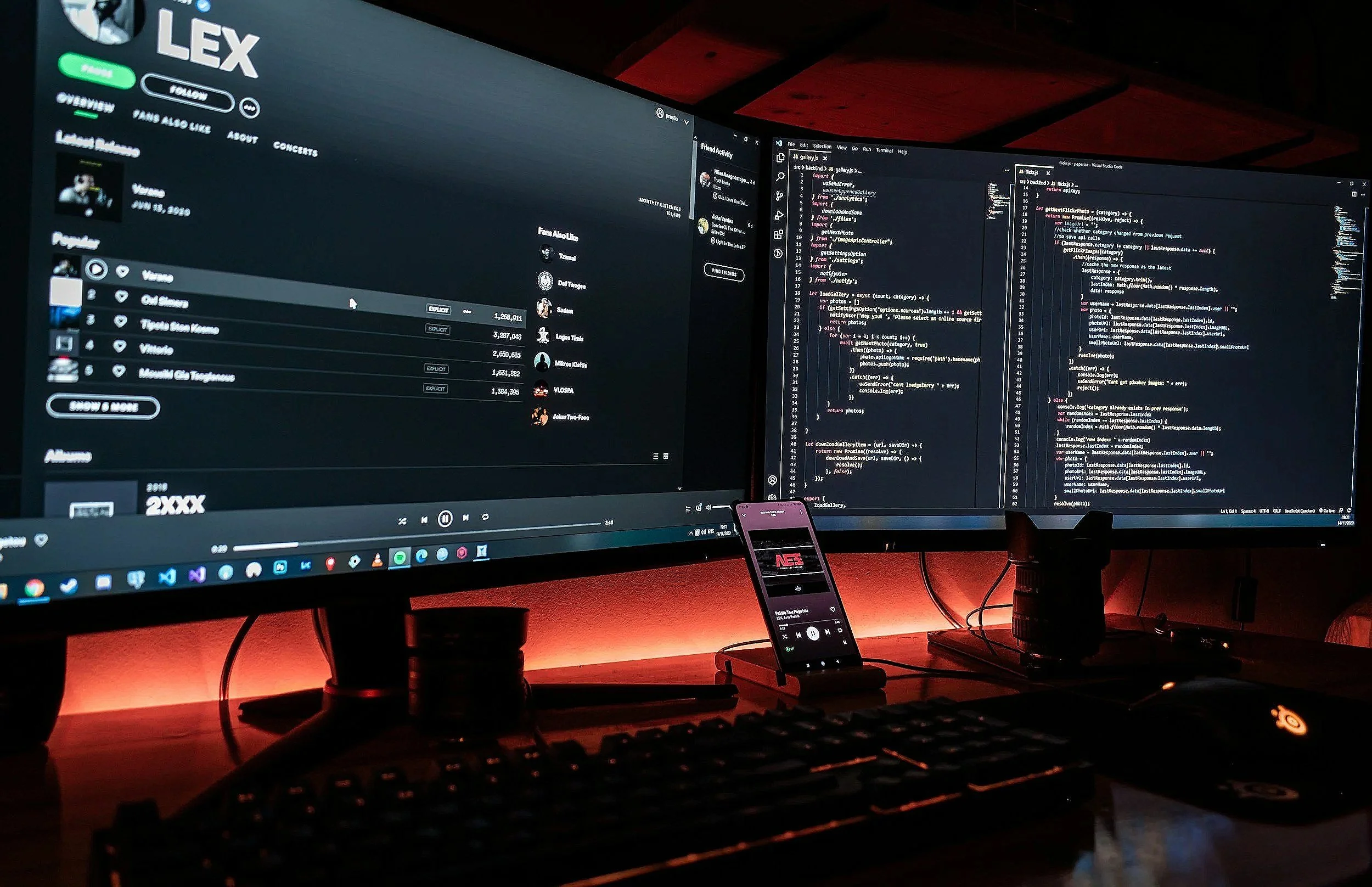Two large computer monitors on a desk, with one displaying a music streaming app and the other showing code in a code editor. There's a keyboard, mouse, a smartphone in a stand, and various cables and black cups on the desk.