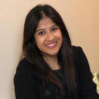 A woman with long dark hair smiling, sitting indoors against a beige wall.