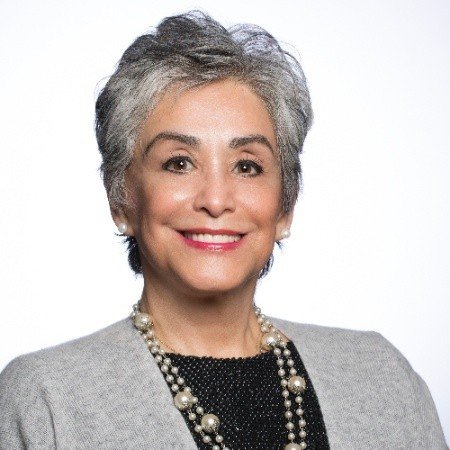 Smiling person with short gray hair, wearing a gray blazer and beaded necklace.