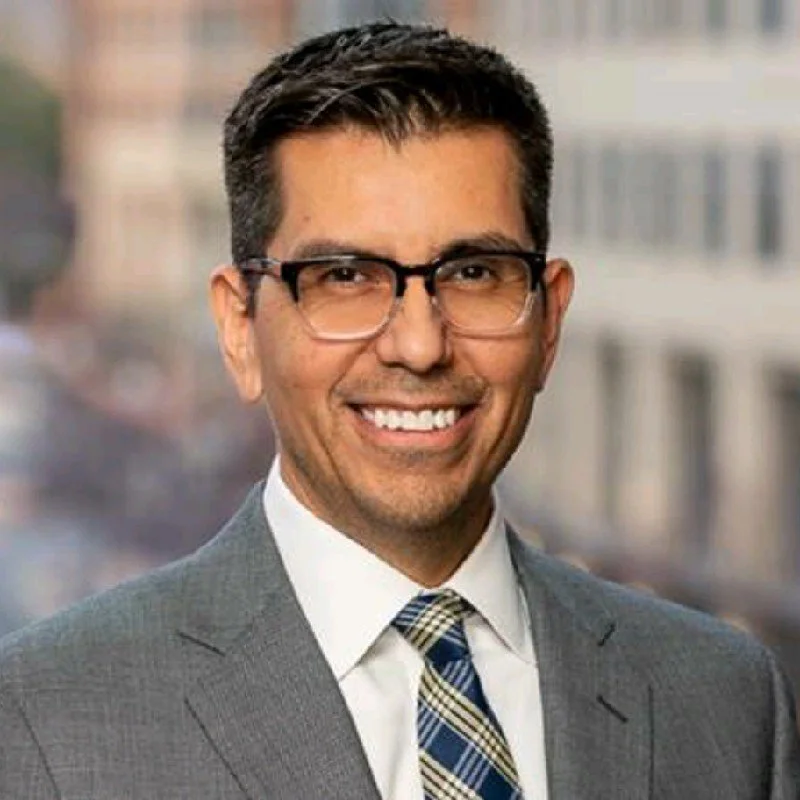 Man in glasses wearing a gray suit with striped tie, smiling, in an urban setting.