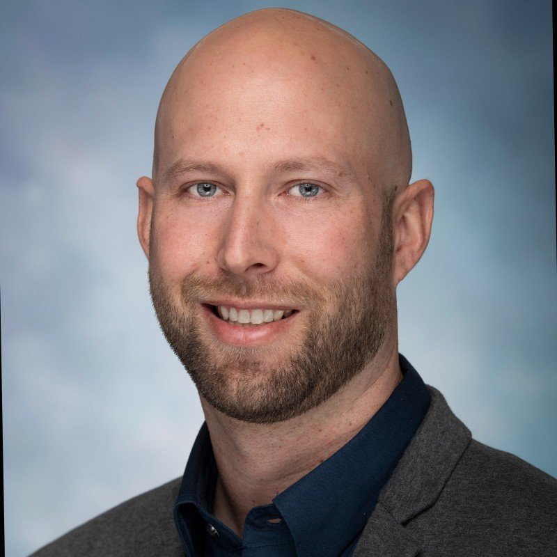 A bald man with a beard, smiling, wearing a dark blue shirt and a gray blazer, against a light, cloudy sky background.