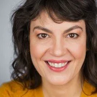 A person with wavy dark hair smiling, wearing a yellow top, against a neutral background.