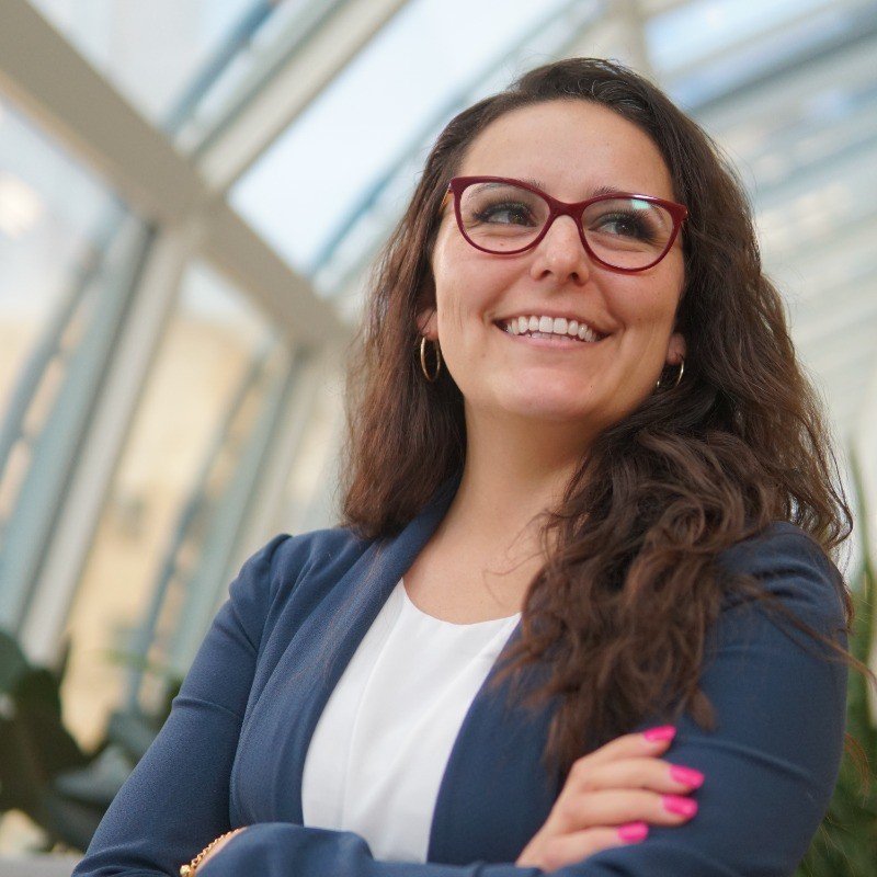 A confident woman with long curly brown hair and glasses, smiling and looking to the side, wearing a navy blazer and a white top inside a glass building.