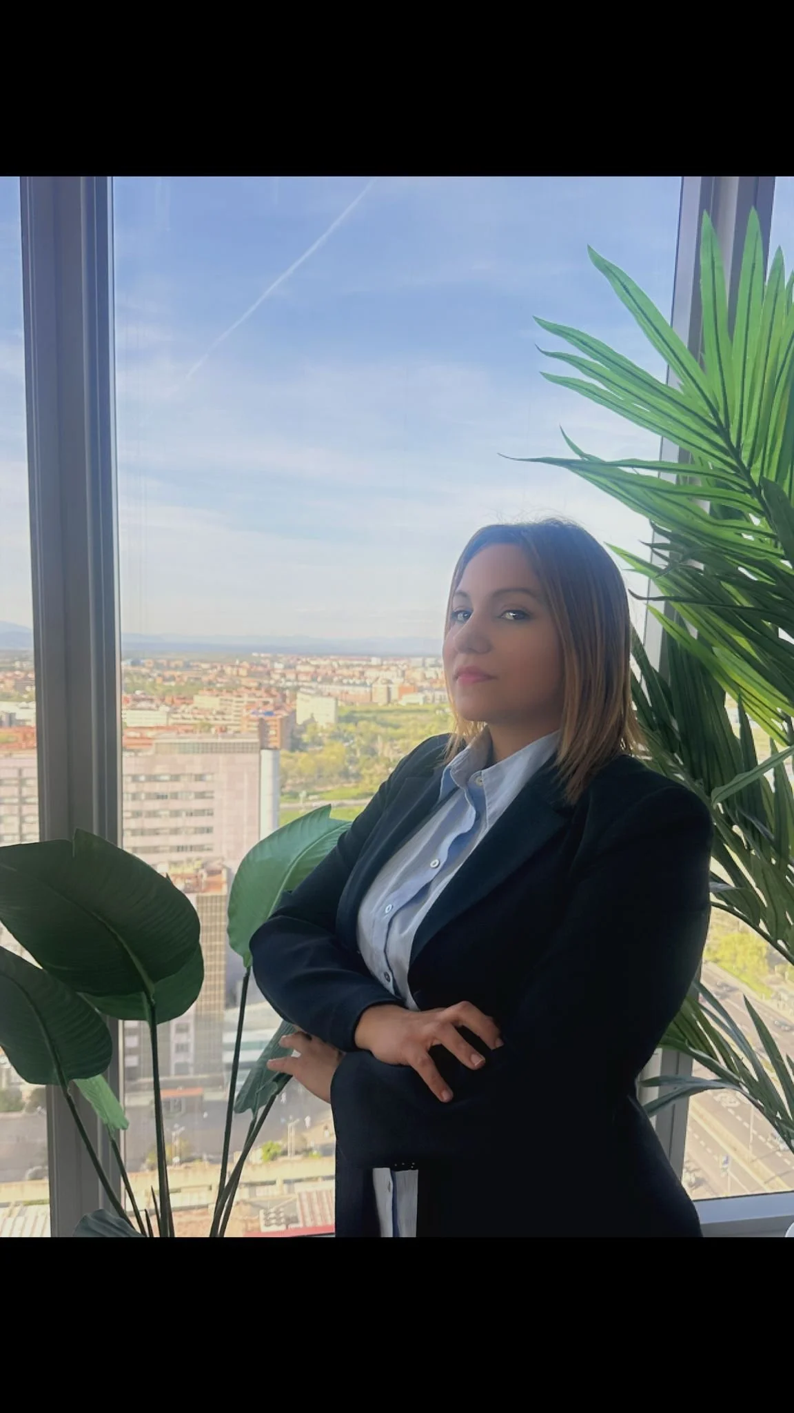 A woman in a black blazer and white shirt standing inside a modern office building with large windows, overlooking a cityscape and blue sky, surrounded by green indoor plants.