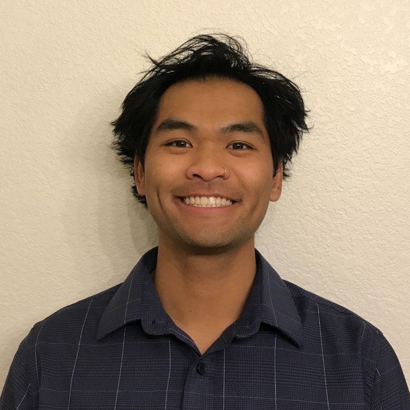 Smiling person with short black hair wearing a dark blue collared shirt, against a light-colored wall.