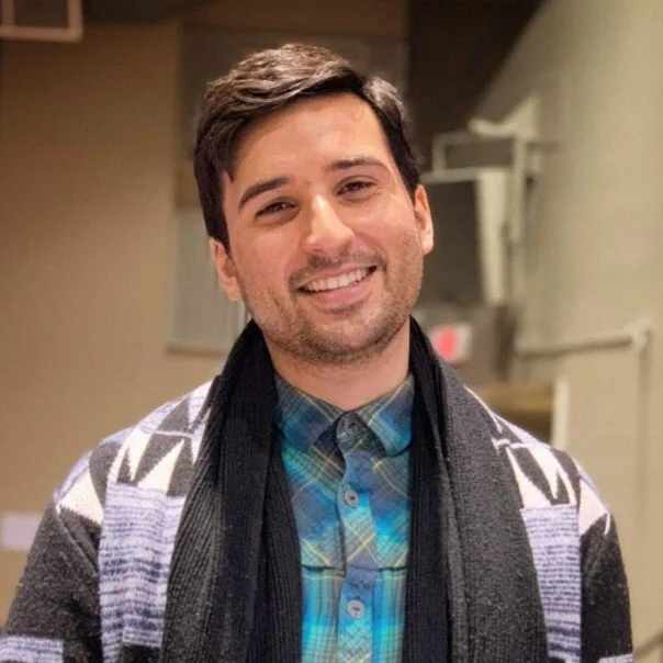A smiling man with dark hair and facial hair wearing a patterned cardigan over a blue and green plaid shirt.