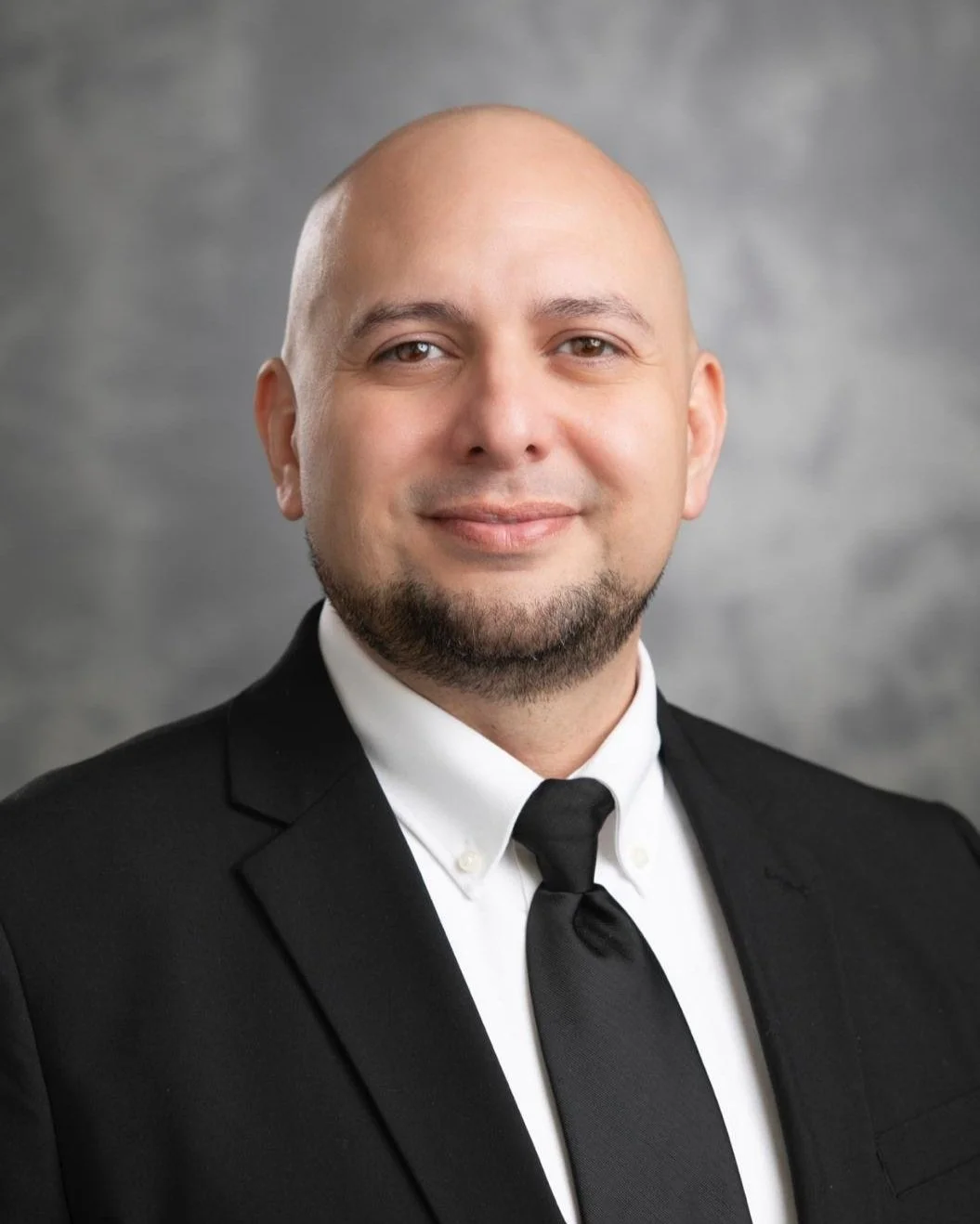 A bald man with a beard in a black suit and tie smiling in a professional portrait.