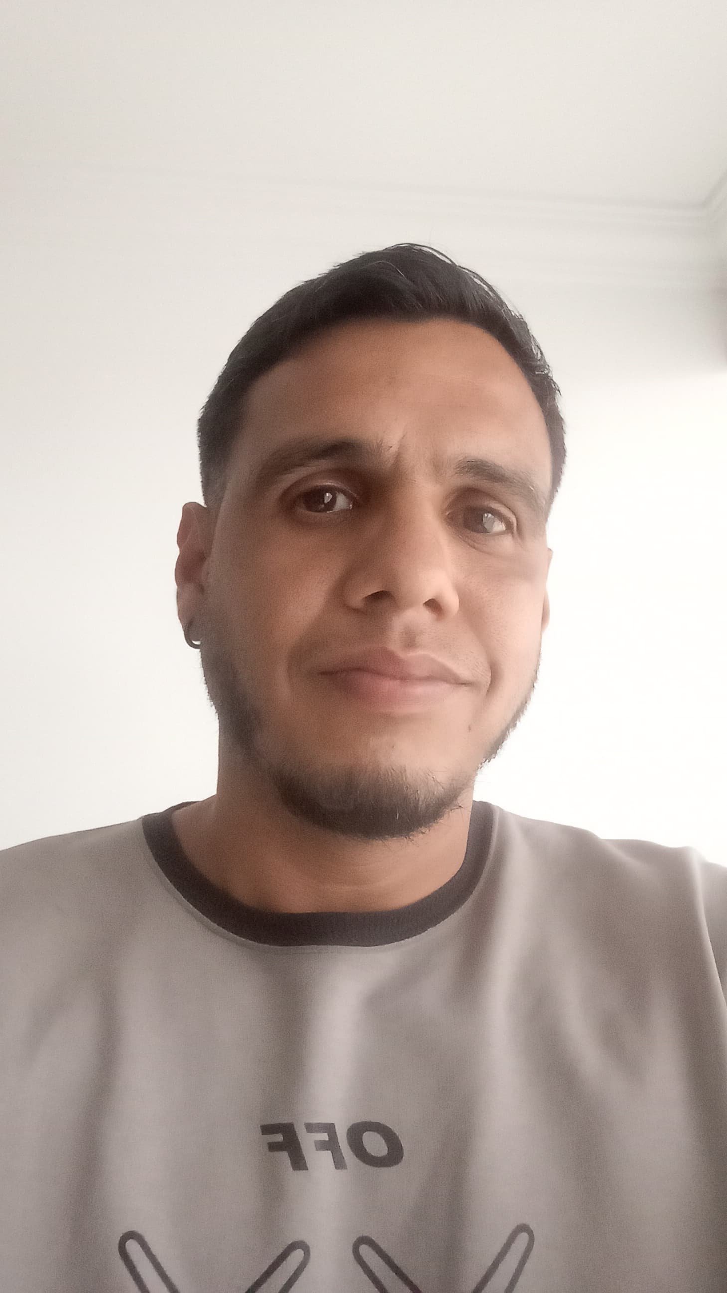 A man with short dark hair, a beard, and earrings taking a selfie indoors against a plain white wall. He is wearing a light-colored shirt with dark trim and some text and graphics on the front.