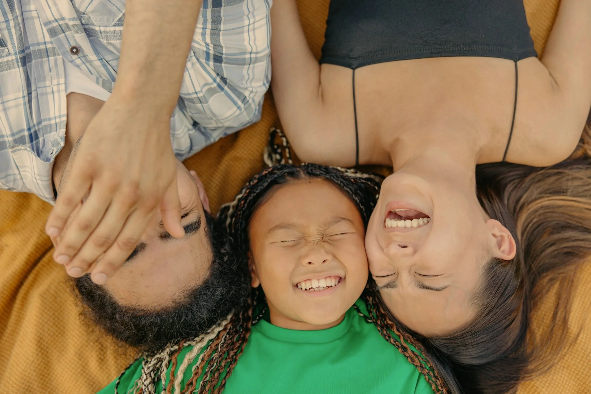 Une photo joyeuse et spontanée d'une jeune fille (4-6 ans) en train de rire aux éclats avec son père et sa mère. Ils sont proches, peut-être en train de jouer ou de partager un moment drôle. L'ambiance est lumineuse, chaleureuse et émotionnelle.