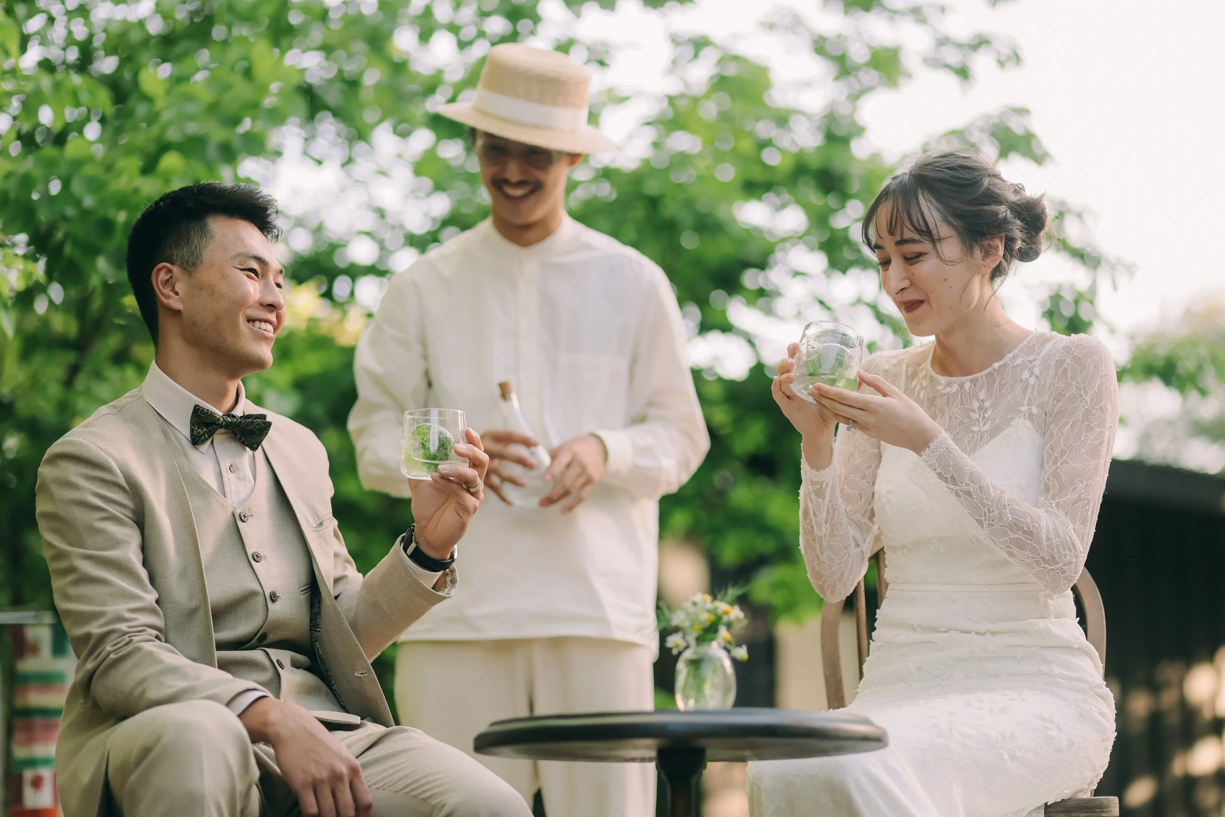 屋外のテーブルで、ボトルを手にしたスタッフとドリンクを試飲する新郎新婦