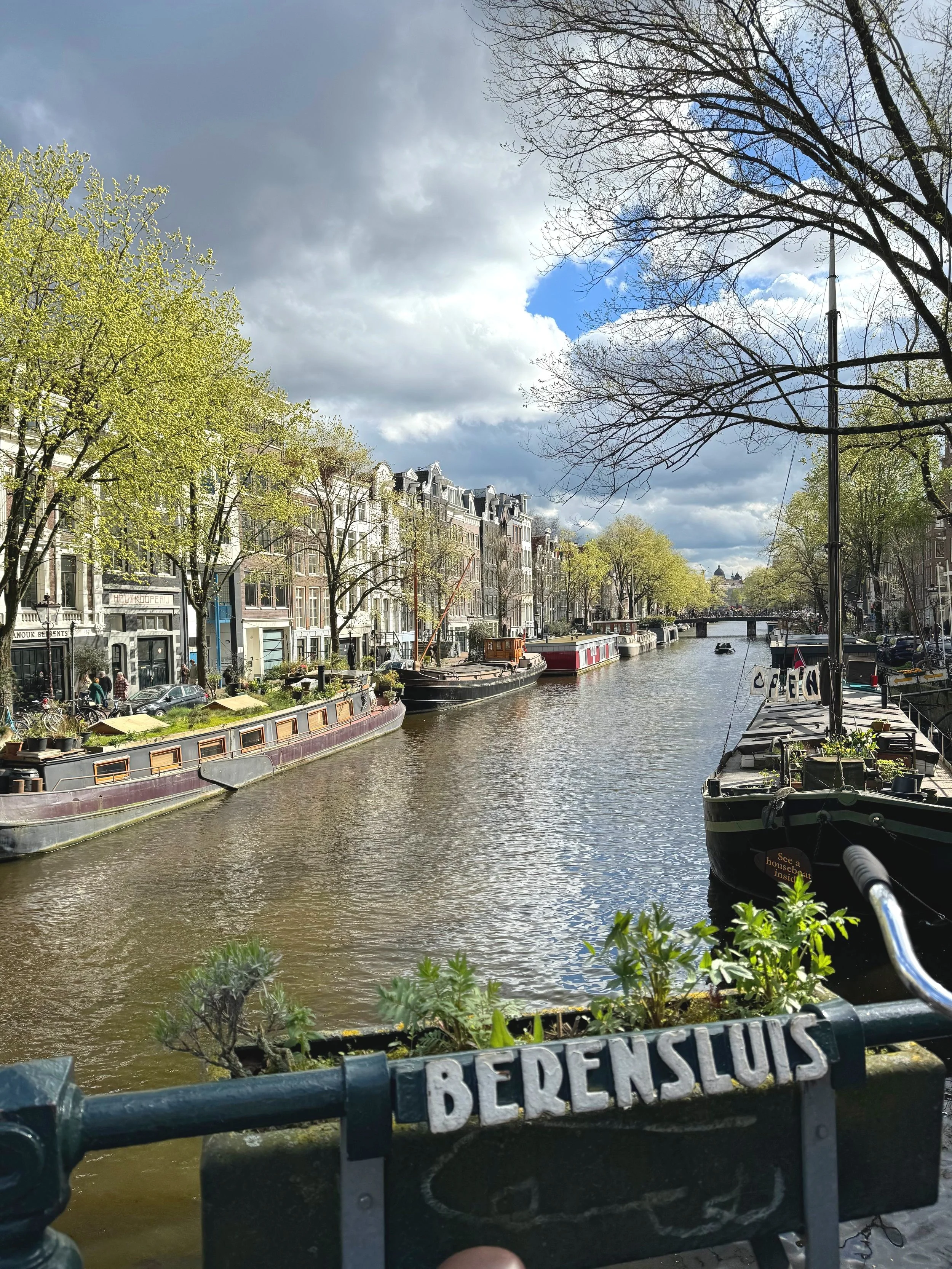 View of a canal in Amsterdam with boats, trees, historic buildings lining the water, and a partly cloudy sky.