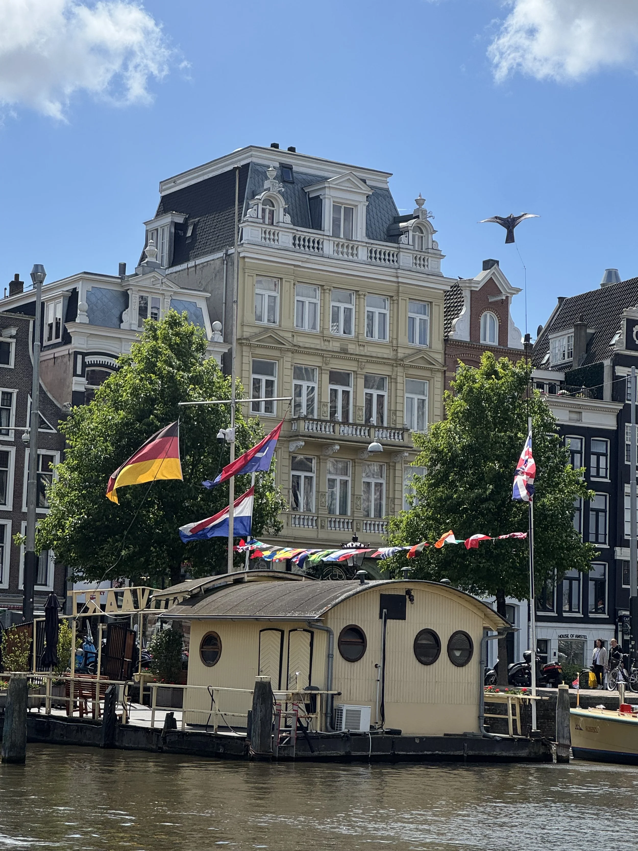 An Amsterdam houseboat floating on the water with various national flags and bunting, with historic Dutch buildings and a flying bird in the background under a blue sky.