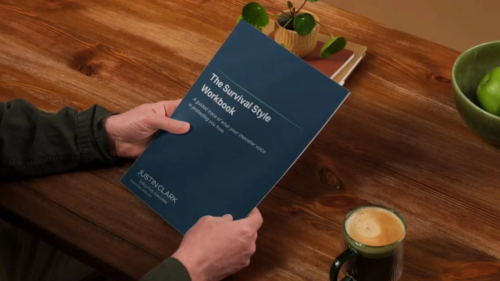Person holding a book titled "The Survival Style Workbook" with a potted plant, a bowl of apples, and a glass of coffee on a wooden table.