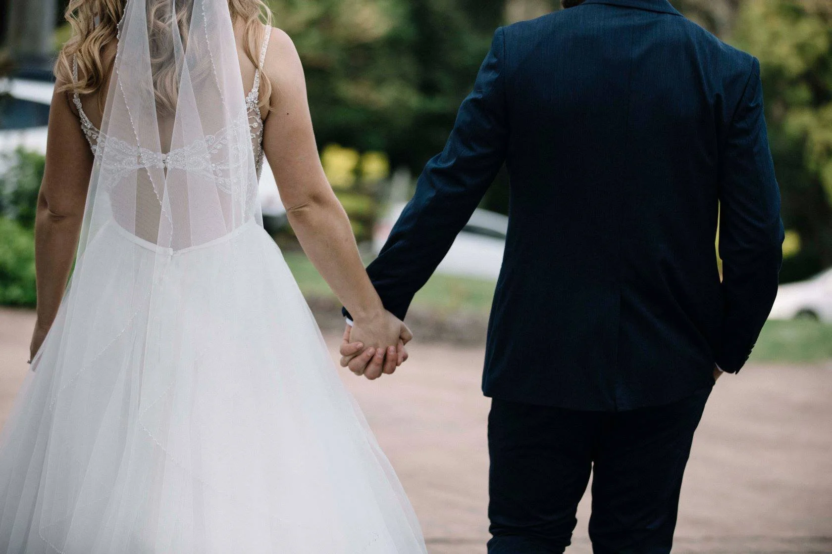 A bride in a white wedding dress and veil holding hands with a groom in a dark suit walking outdoors.