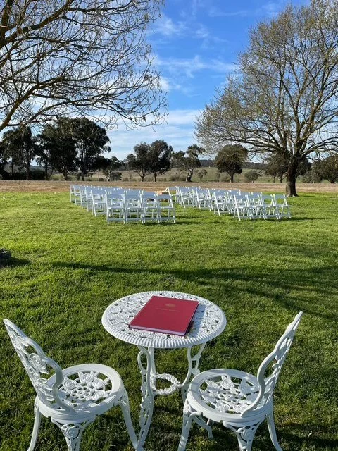Outdoor wedding setup with white chairs arranged in rows on a grassy field, a small white table with two chairs in the foreground, and a red book on the table, with trees and a blue sky in the background.