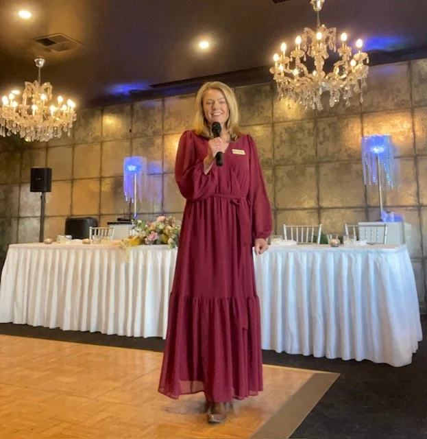 A woman in a long burgundy dress holding a microphone and smiling at a formal event in a decorated banquet hall.