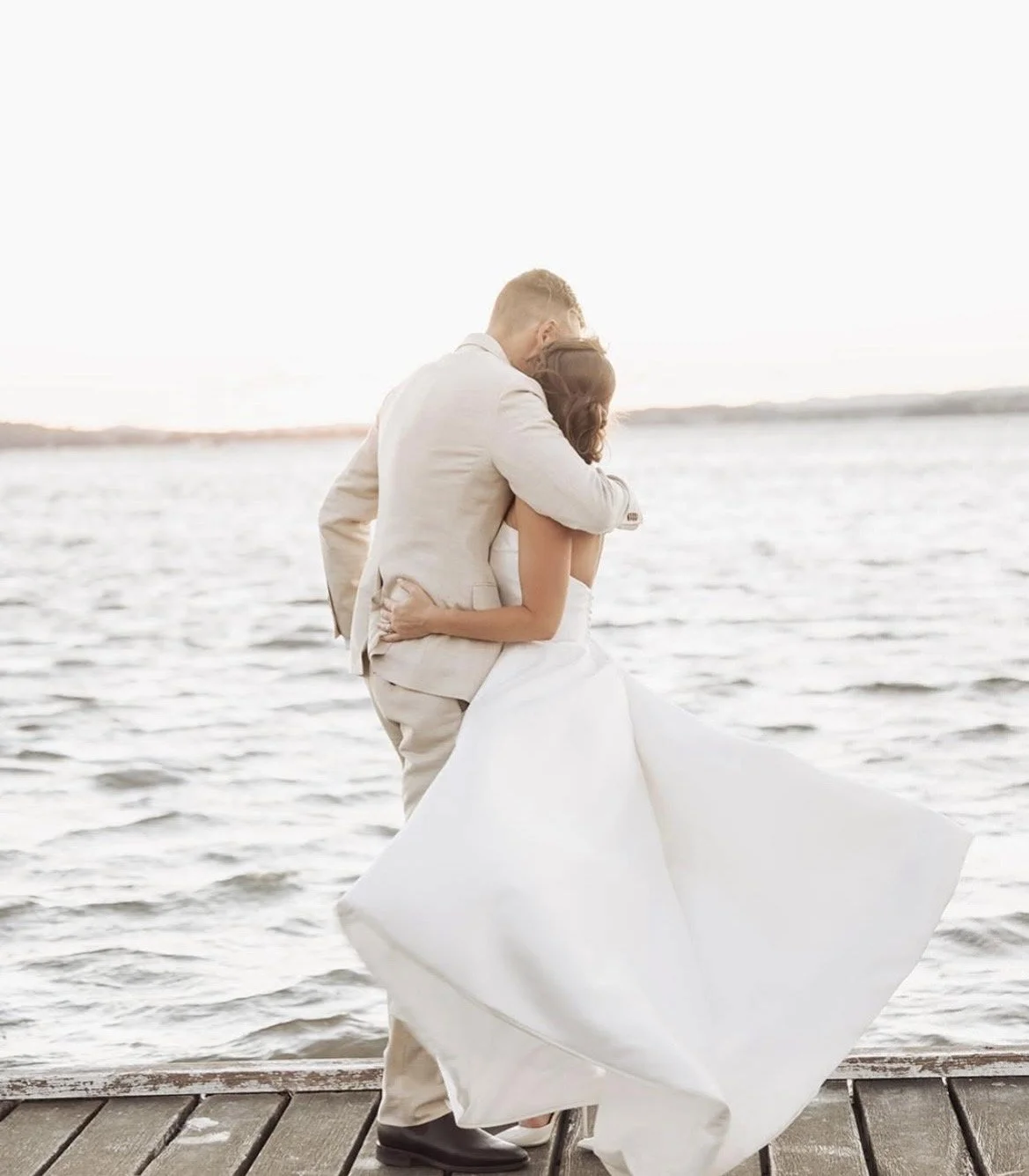 A bride and groom embracing by the water on a dock during sunset.