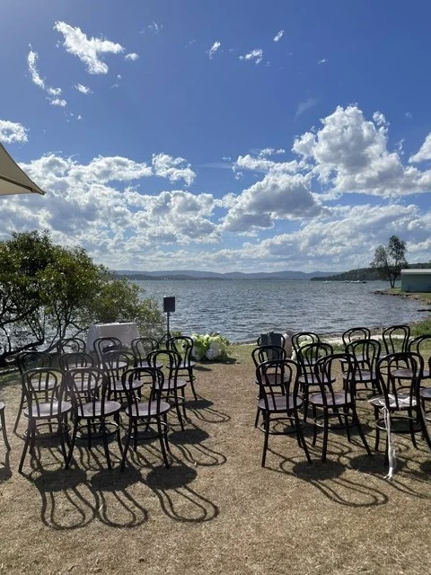 Empty black chairs arranged outdoors near a lake, with a cloudy blue sky overhead.