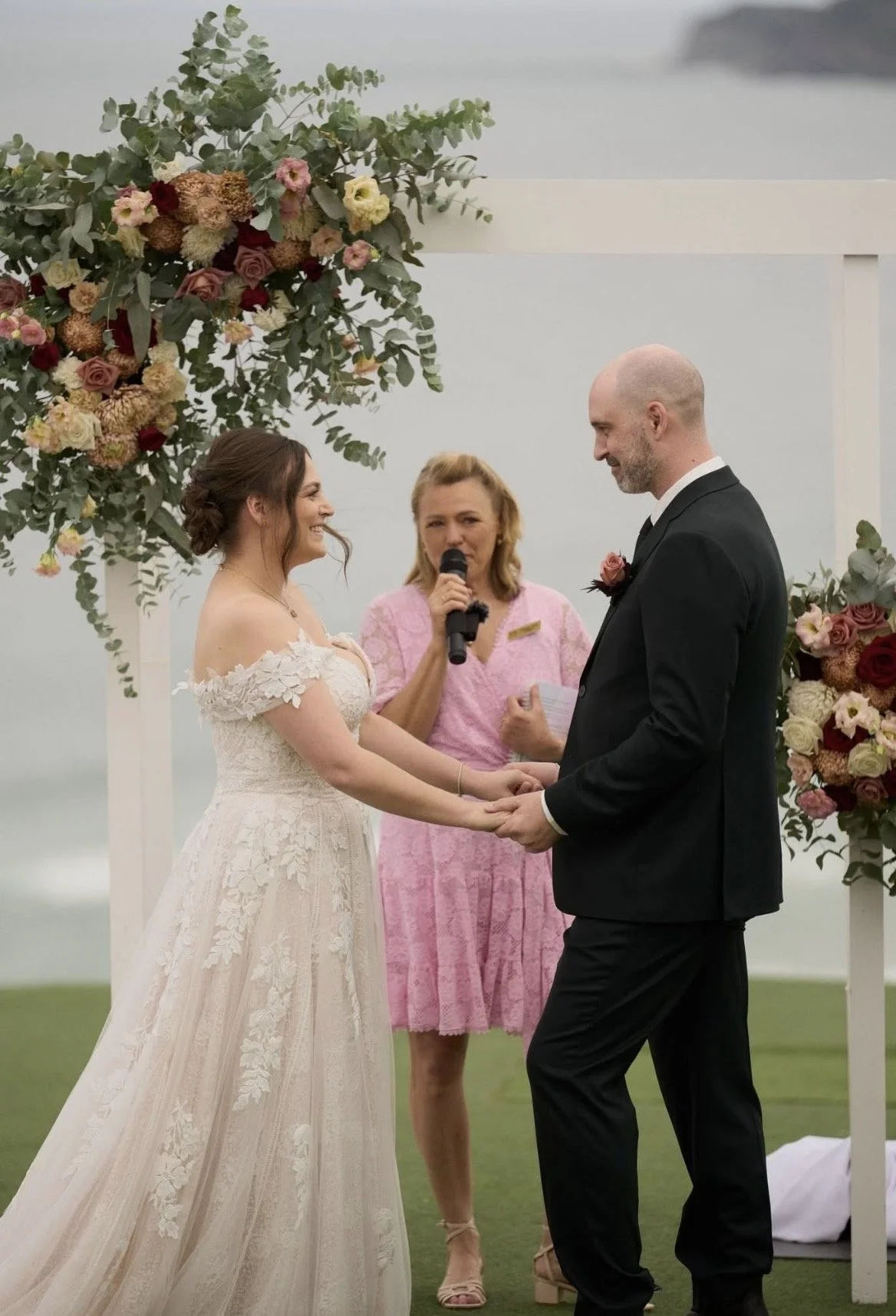 A wedding ceremony with a bride and groom holding hands, exchanging vows, standing under a floral arch, with an officiant in a pink dress holding a microphone.