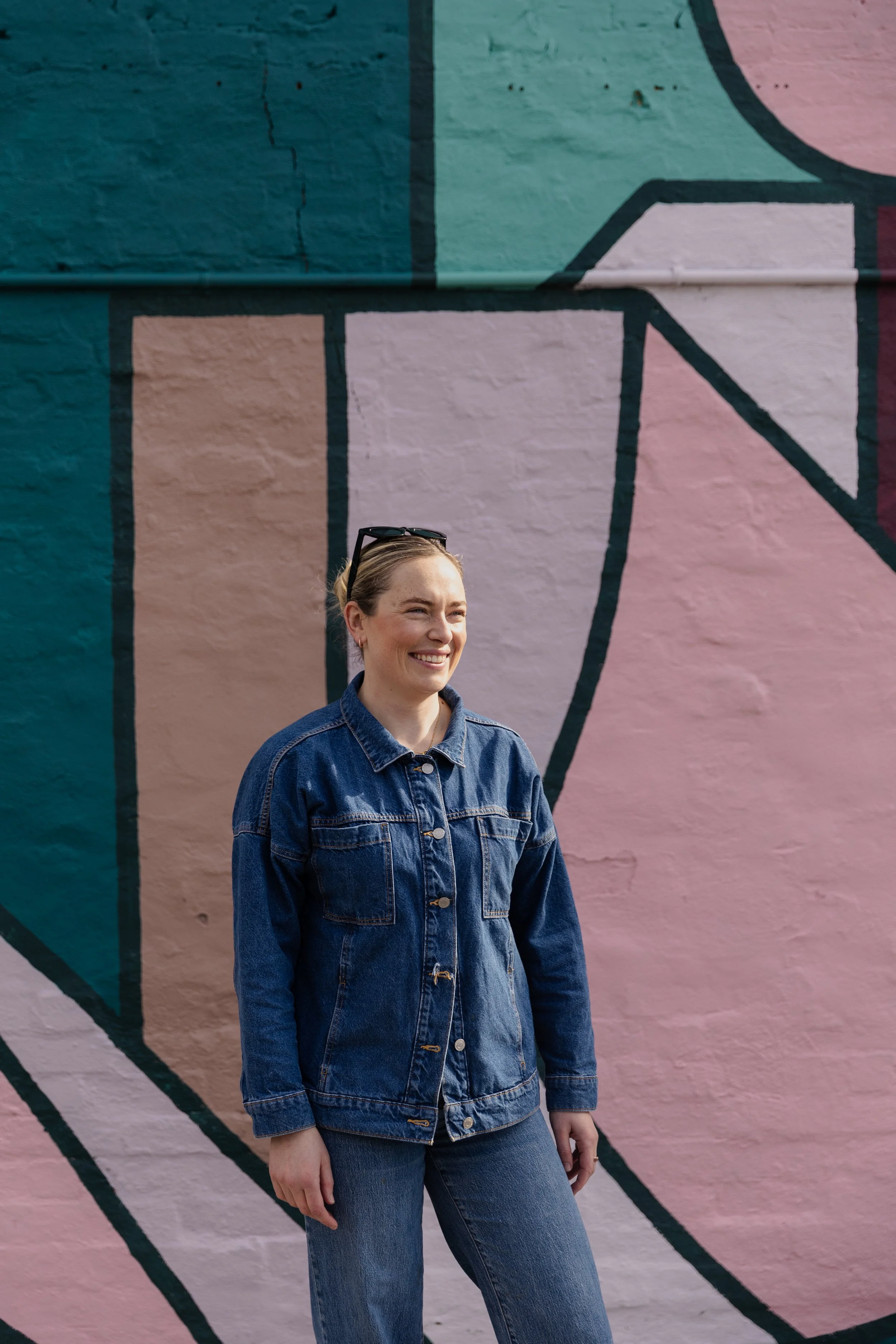 Imogen, a woman, stands smiling in front of a colorful mural painted on a brick wall, wearing a denim jacket and jeans with sunglasses resting on her head.