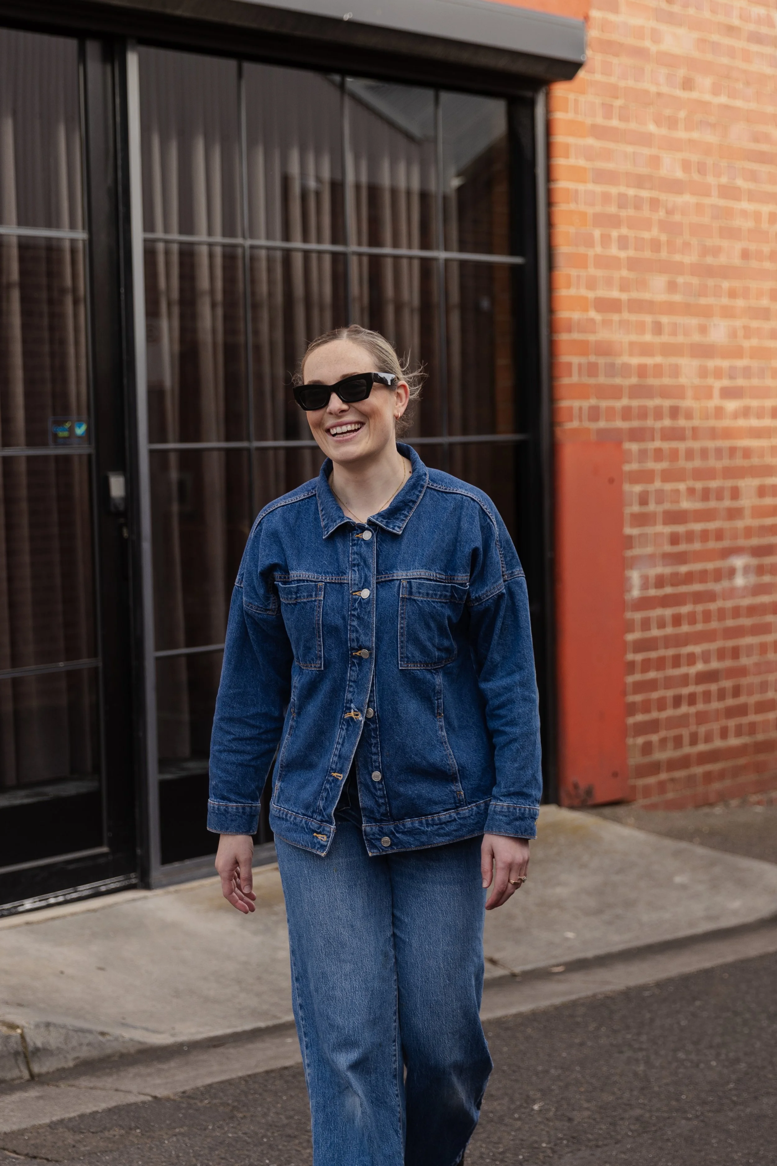 Imogen, a woman, walking outdoors in front of a brick building, wearing sunglasses, a denim jacket, and jeans, smiling.